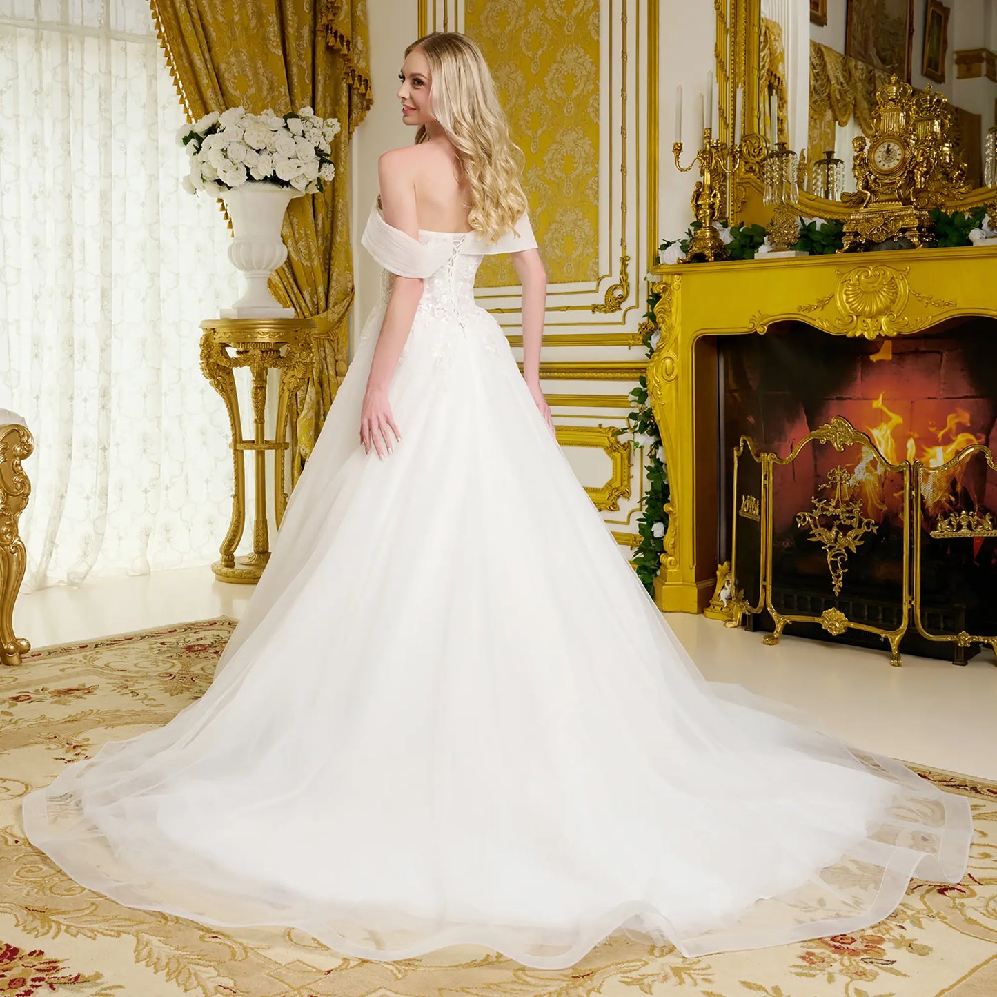 Woman in a white wedding dress standing in an opulent room with gold decor and a fireplace.