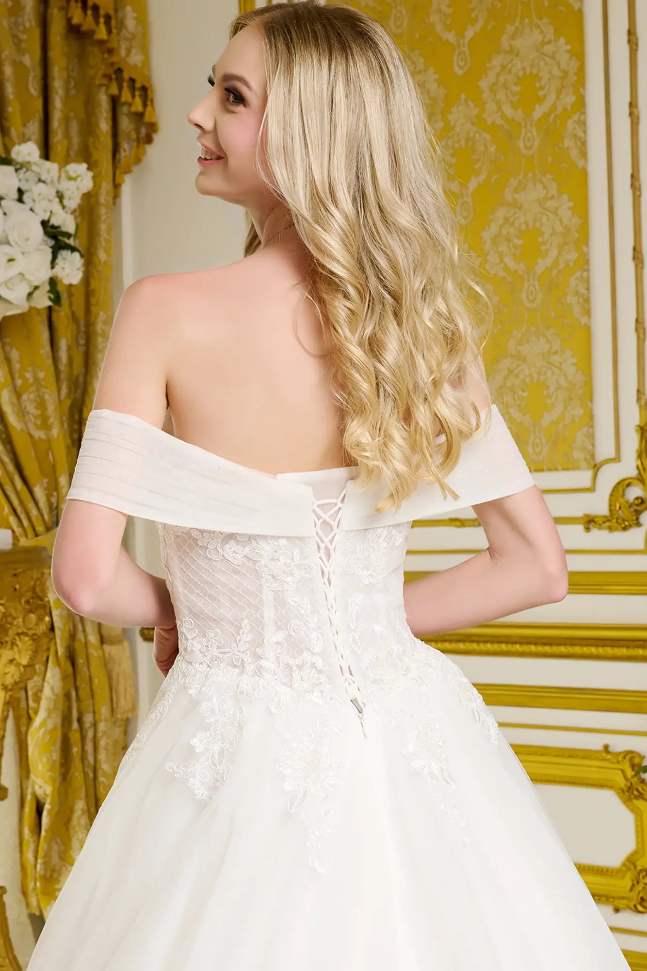 Woman in a white off-shoulder wedding dress standing in an ornate room with gold accents.