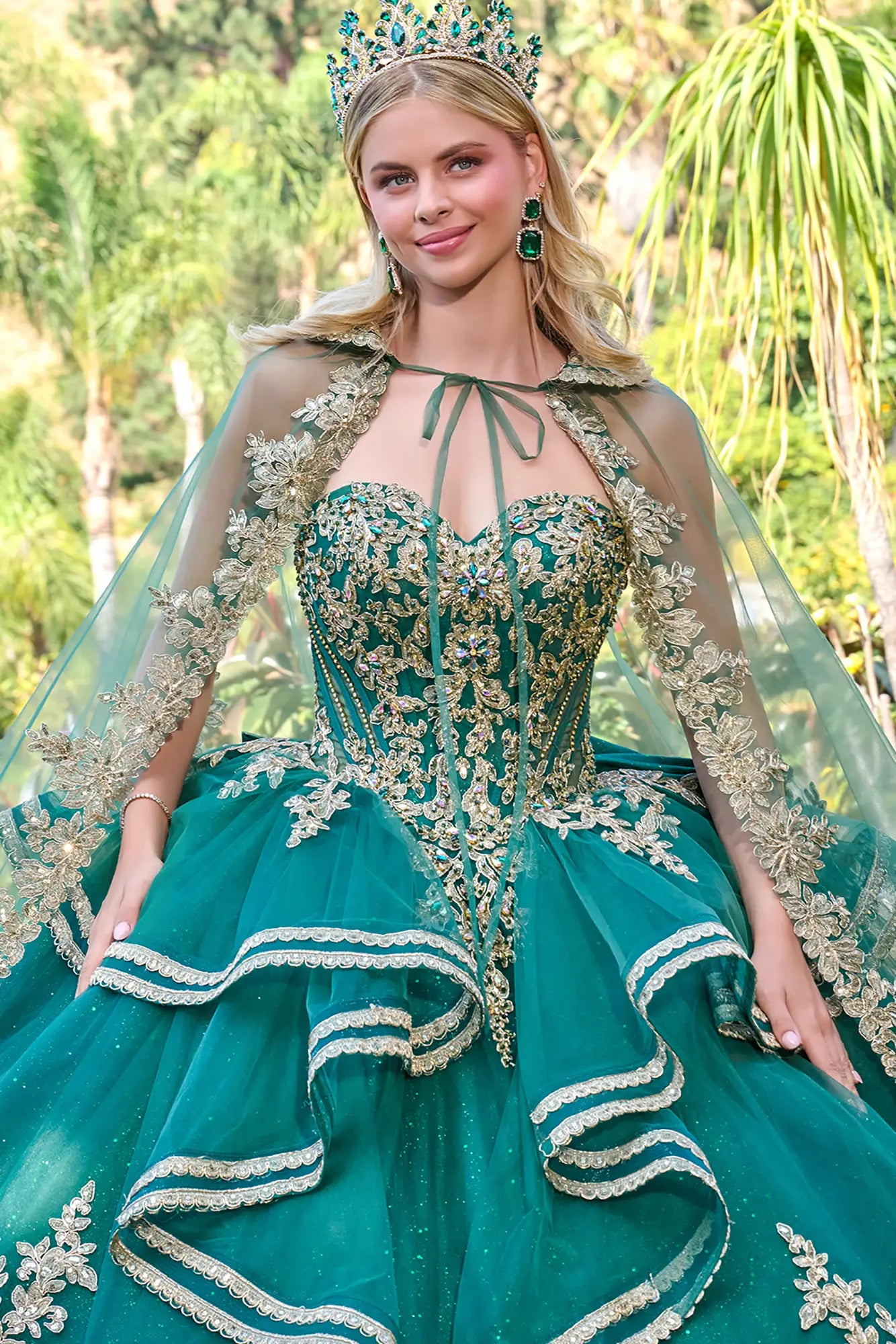 Woman in a green quinceañera dress with a floral cape and tiara, standing outdoors.