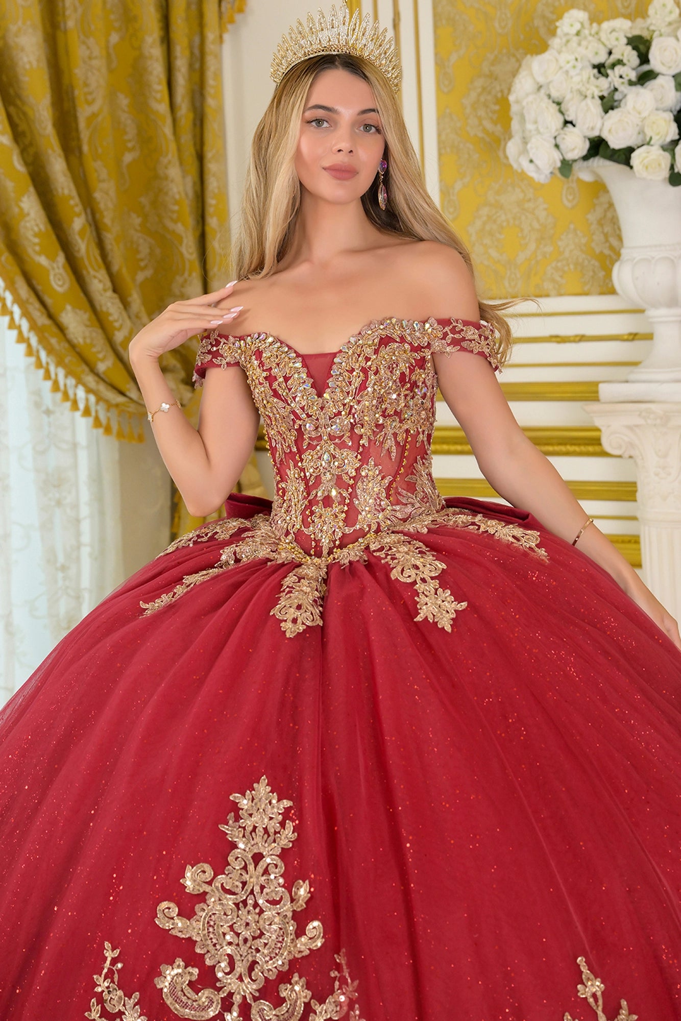 Front view of burgundy quinceañera ball gown featuring off-the-shoulder straps, sheer embroidered corset bodice with gold beadwork, and full glitter tulle skirt