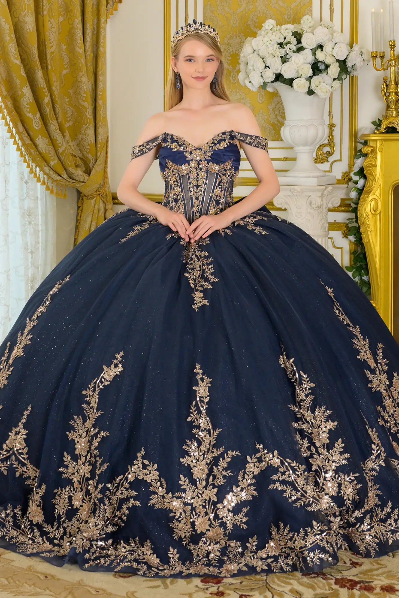 Front view of navy quinceañera ball gown with off-the-shoulder neckline, gold embroidered corset bodice, structured waist, and full glitter tulle skirt in an elegant palace interior