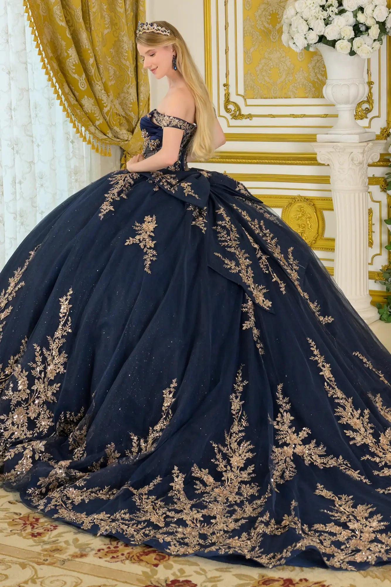 Side back view of navy quinceañera ball gown featuring off-the-shoulder straps, gold embroidery, oversized detachable bow, and voluminous layered tulle skirt