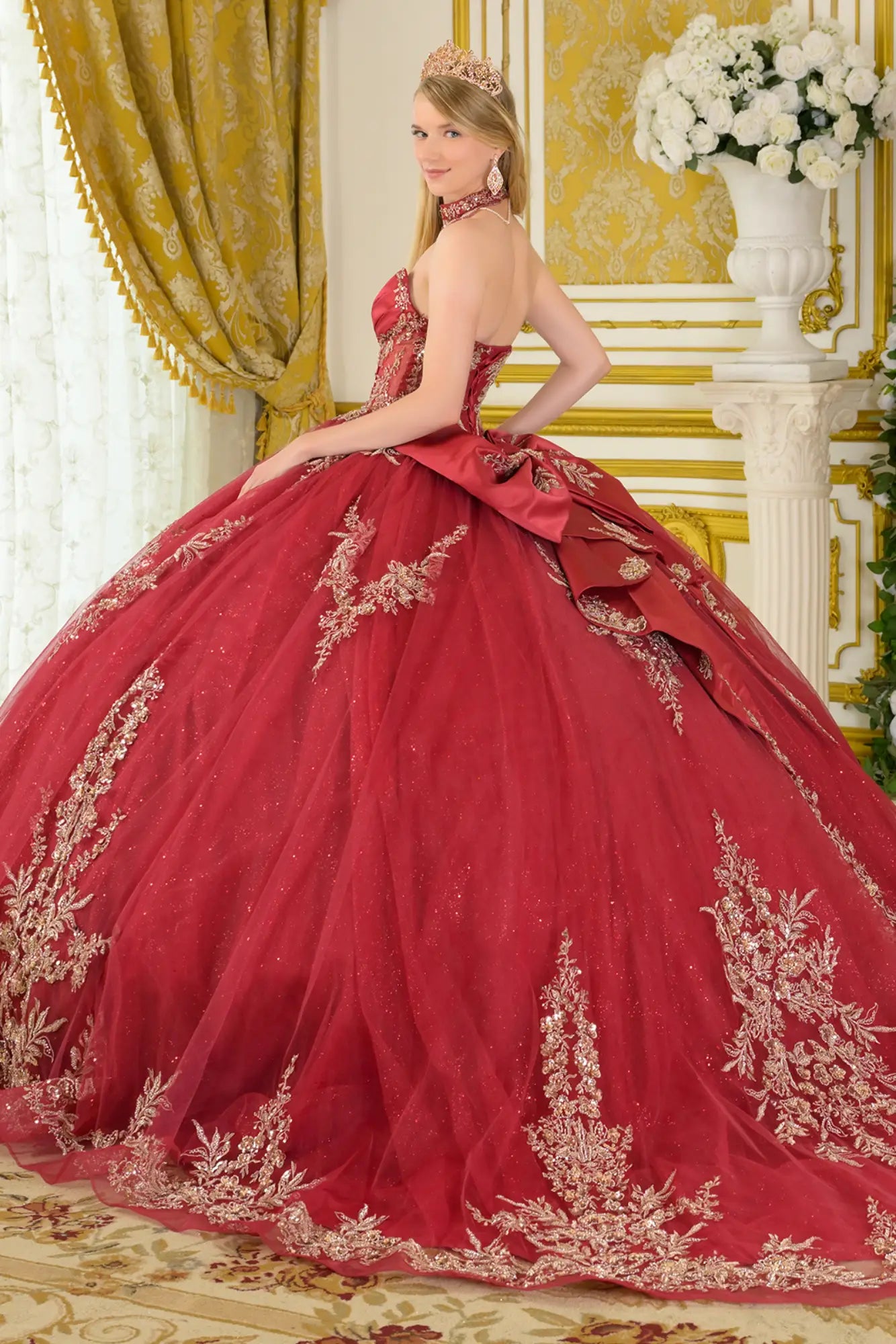 Side back view of burgundy quinceañera ball gown featuring strapless corset, gold embroidery, oversized detachable bow, and full layered tulle skirt