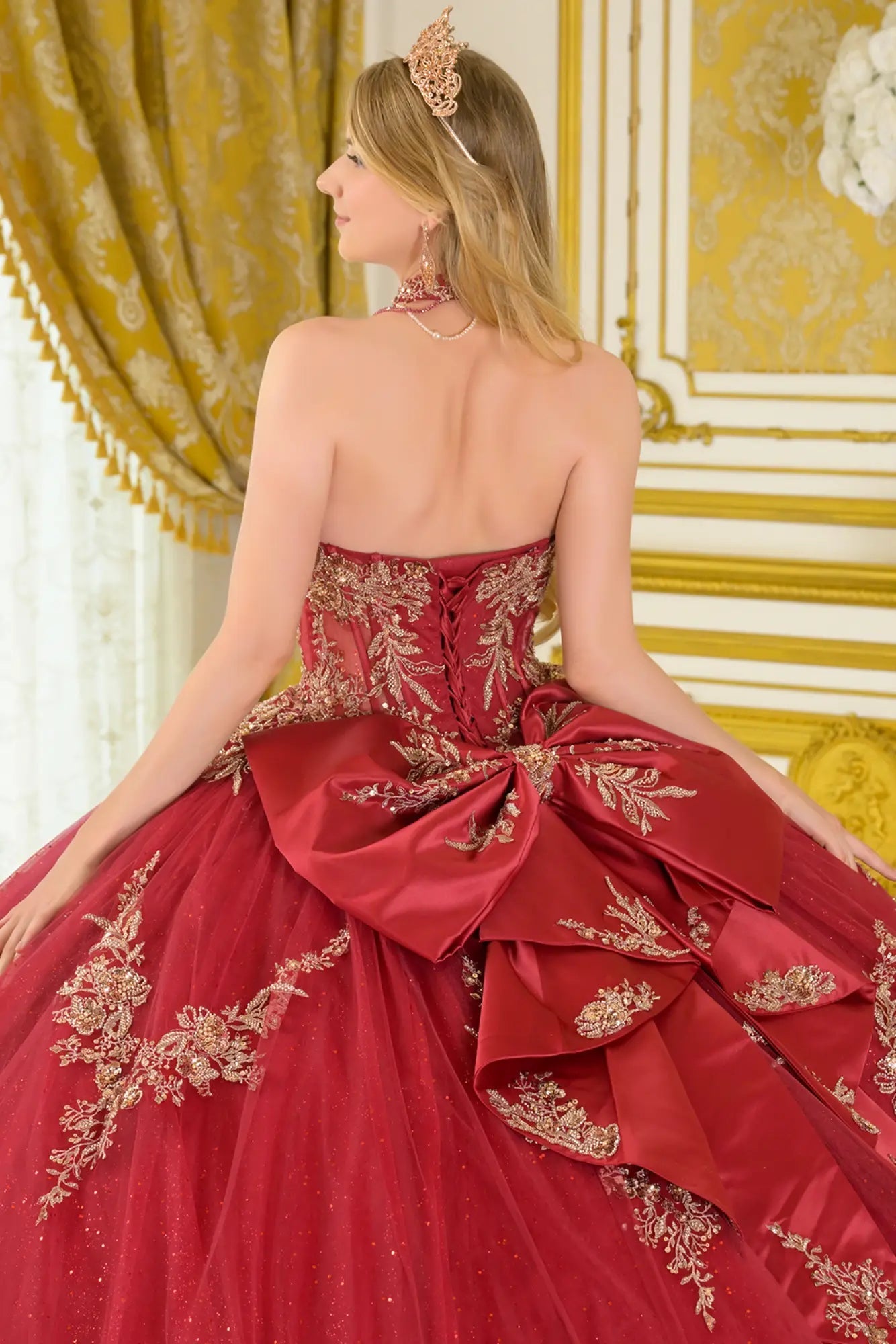 Close-up back view of burgundy quinceañera ball gown with lace-up corset back, gold embroidered detailing, detachable bow, and voluminous tulle skirt