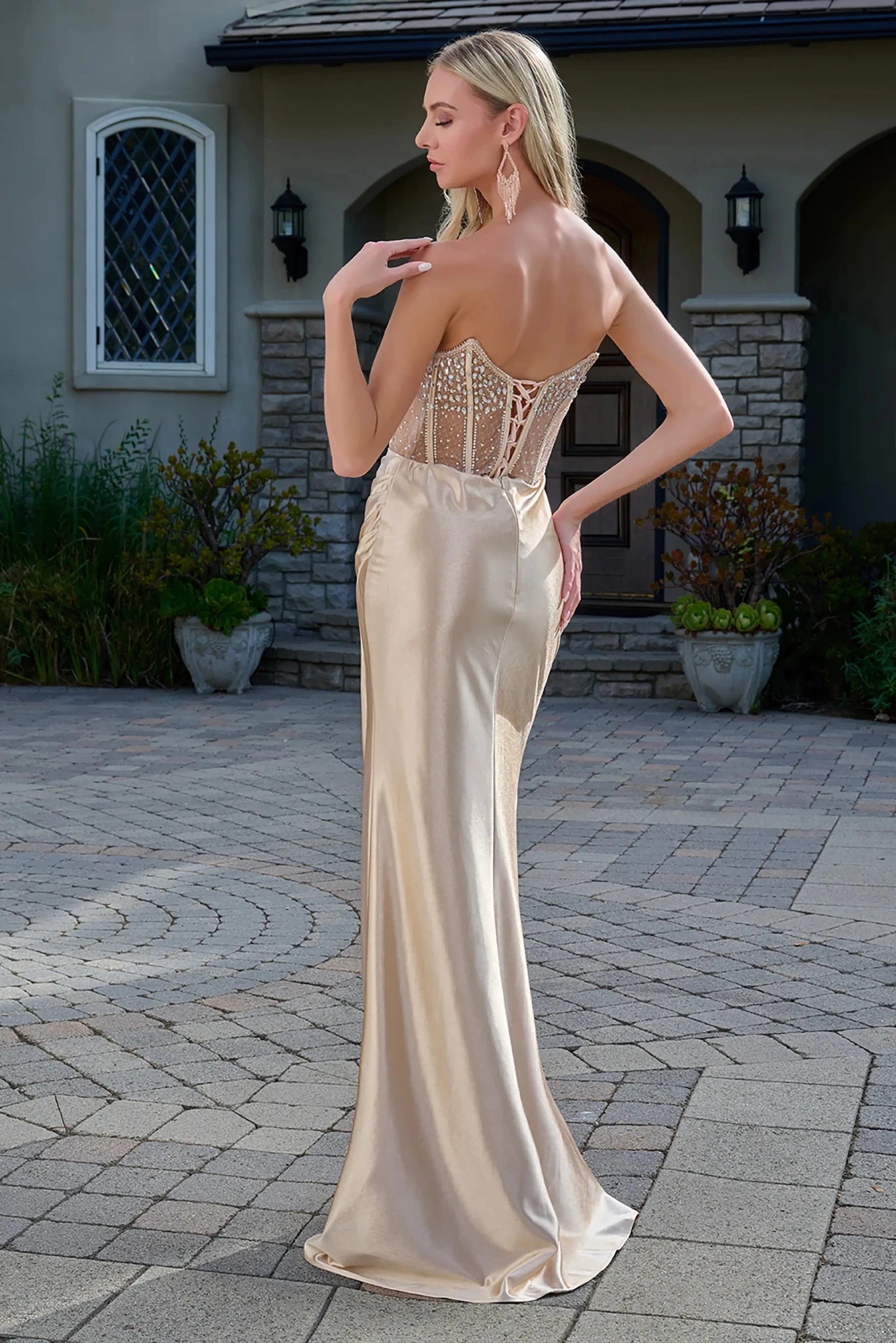 Back full-length view of a champagne strapless prom dress with a sheer beaded corset bodice, boat neckline, fitted satin skirt, and a thigh-high slit in a floor-length design.