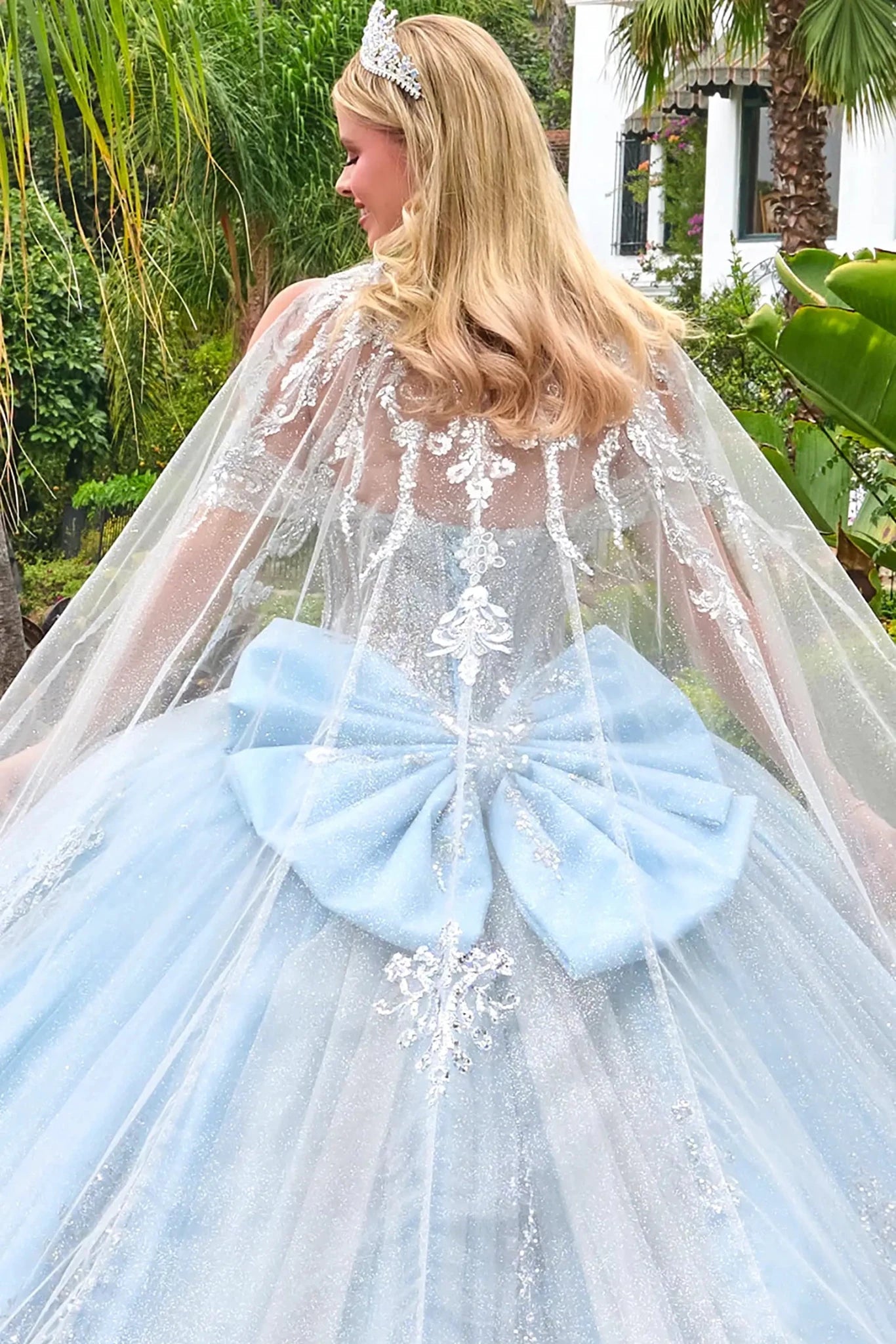 Woman in a light blue dress with a large bow and sheer cape outdoors.
