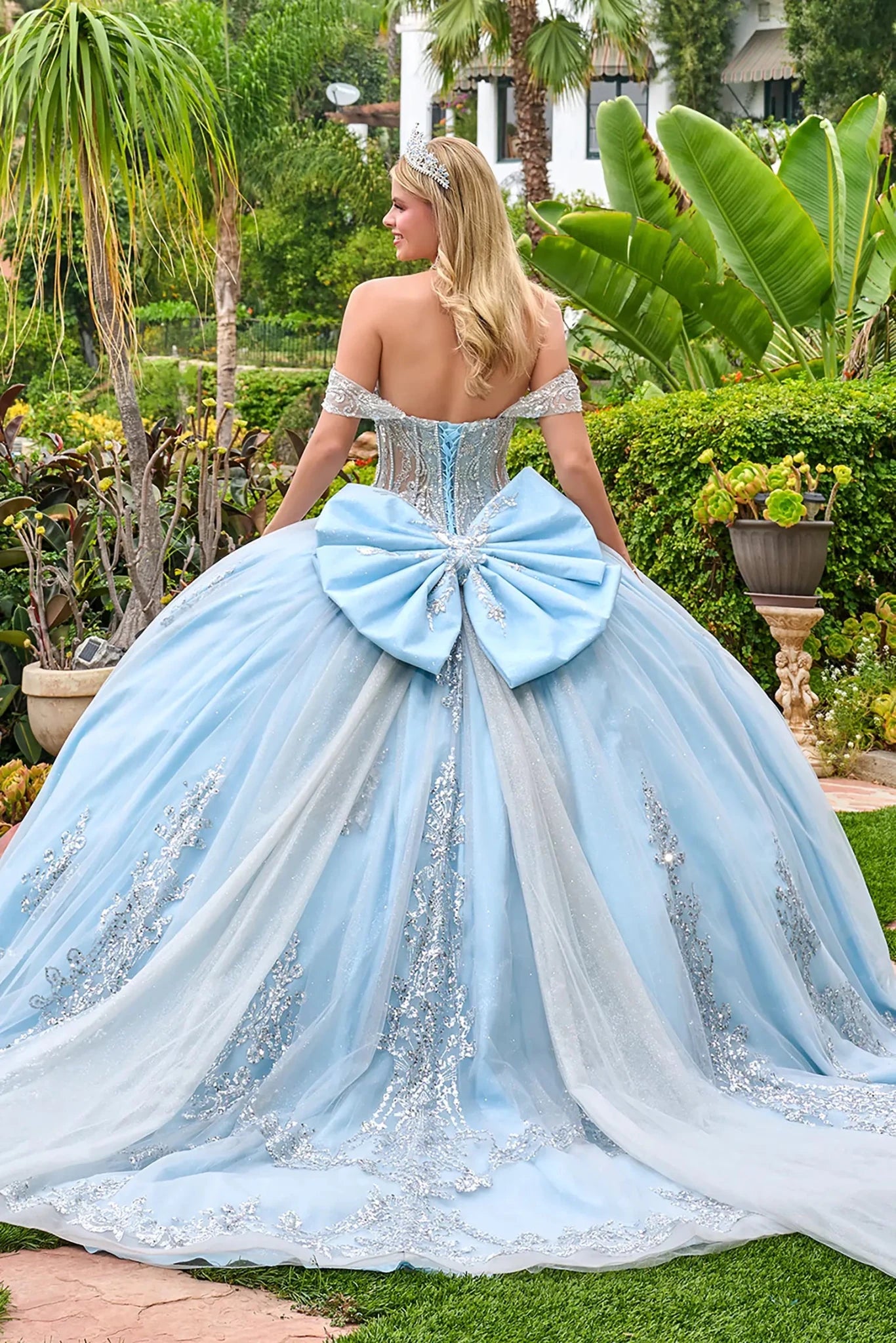 Woman in a light blue and white ball gown with a large bow in an outdoor garden setting.