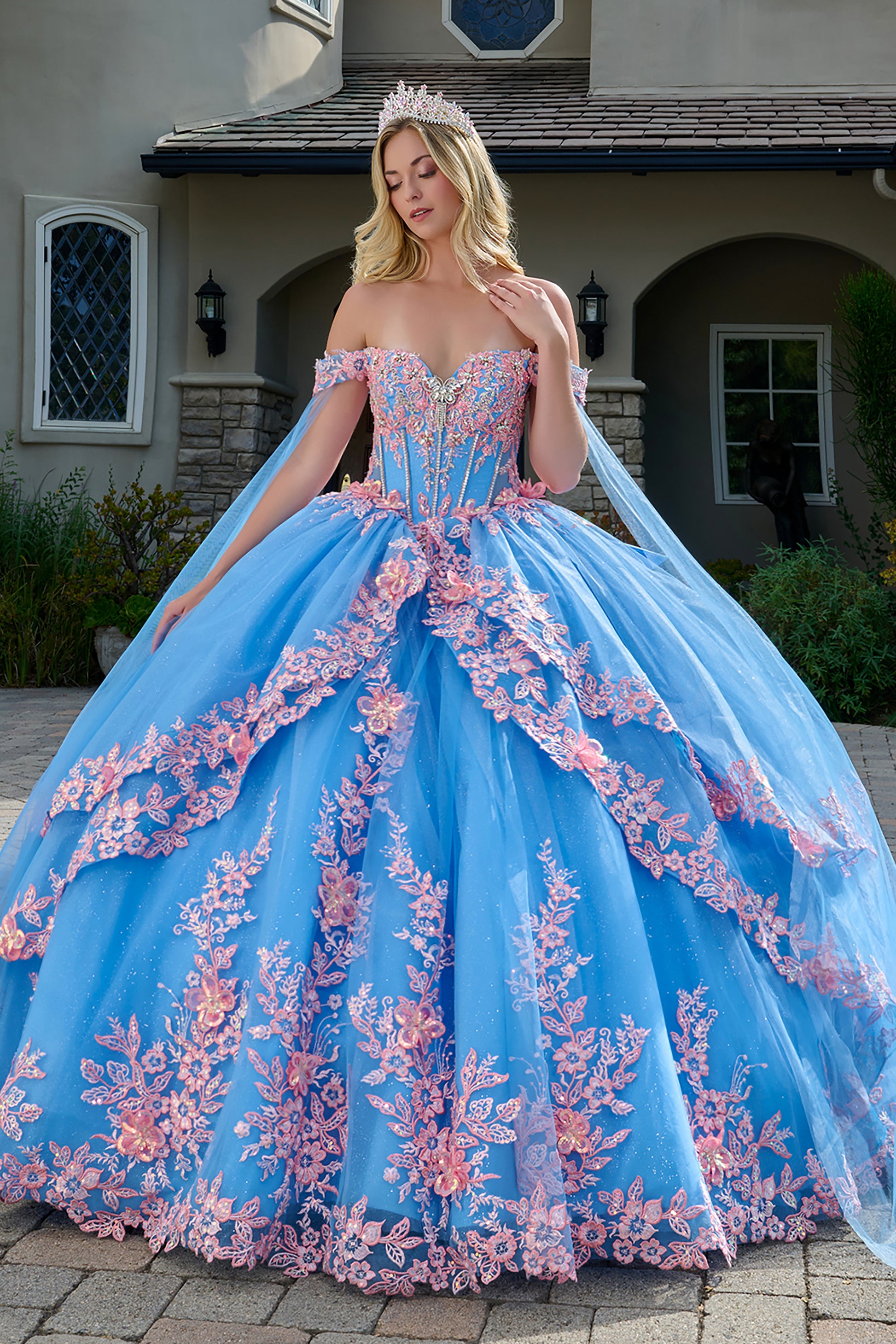 Woman in a blue and pink ball gown with floral embroidery standing in front of a house.