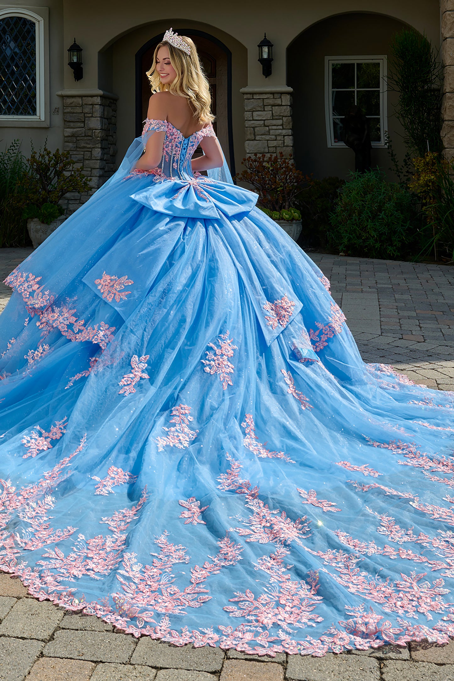 Woman in a blue and pink formal gown with a tiara, standing outside a building.