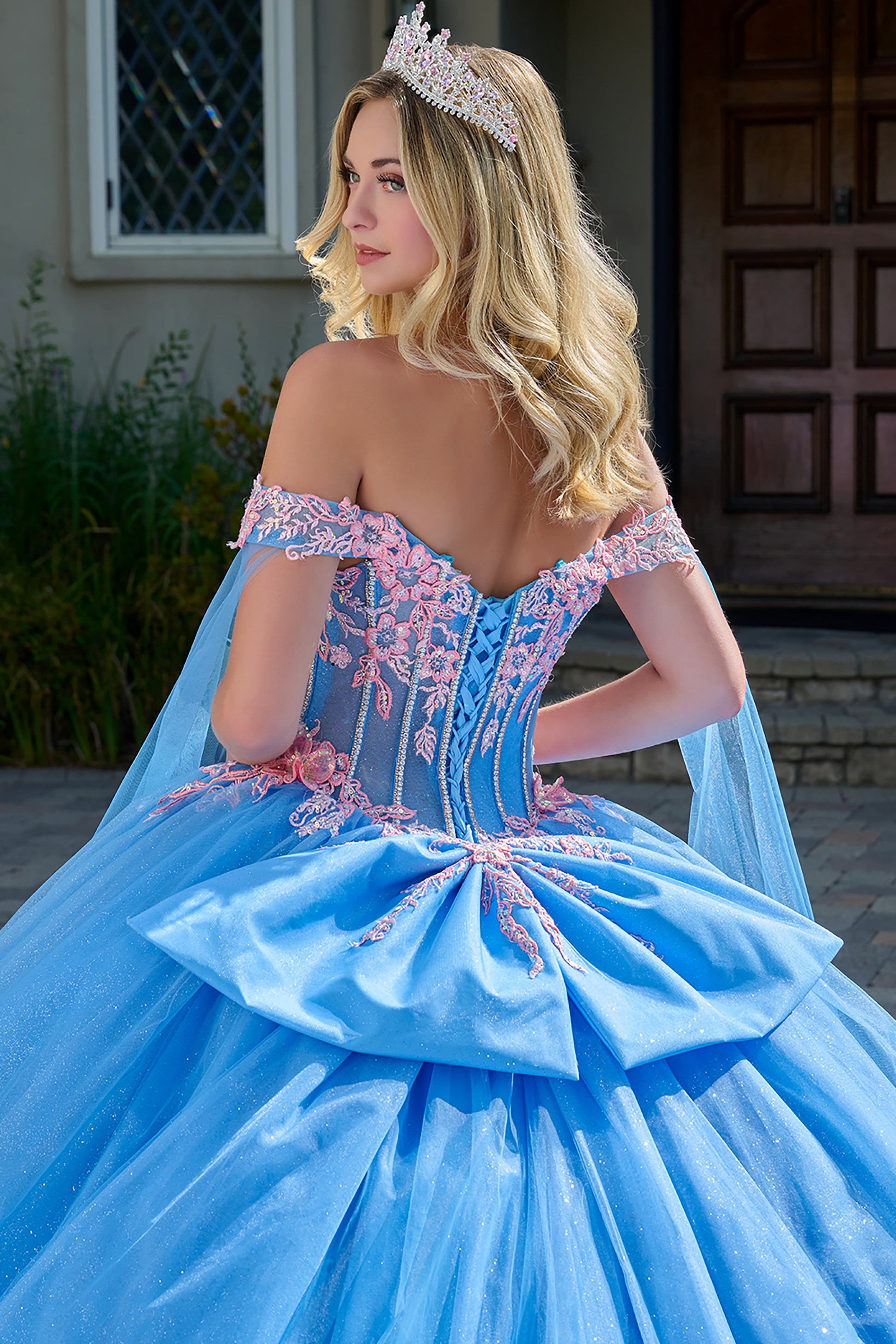 Woman in a blue ball gown with floral details and a tiara, standing outdoors.
