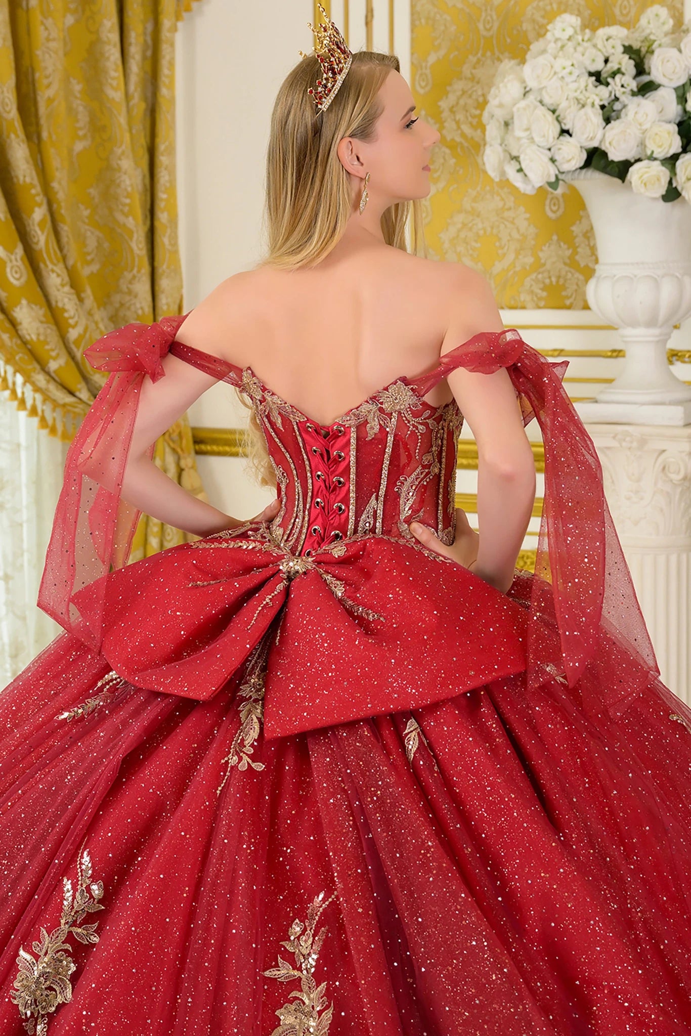 The image shows a woman standing in a luxurious indoor setting decorated with gold-patterned curtains, a white column, and a vase of white flowers. She is wearing an elaborate sparkling red ball gown with a fitted corset-style bodice adorned with intricate gold embellishments.