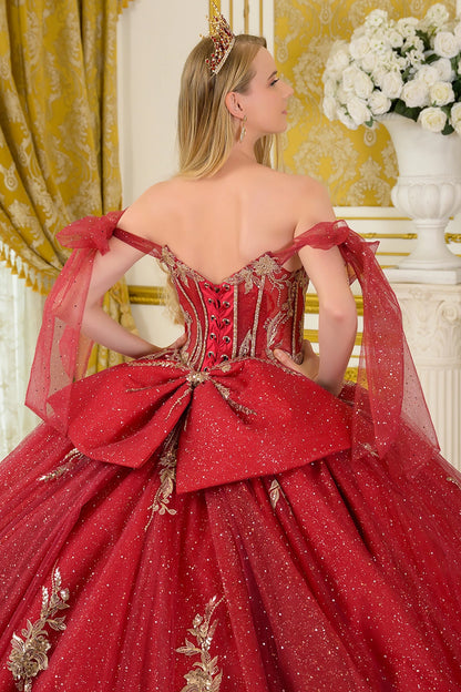 The image shows a woman standing in a luxurious indoor setting decorated with gold-patterned curtains, a white column, and a vase of white flowers. She is wearing an elaborate sparkling red ball gown with a fitted corset-style bodice adorned with intricate gold embellishments.