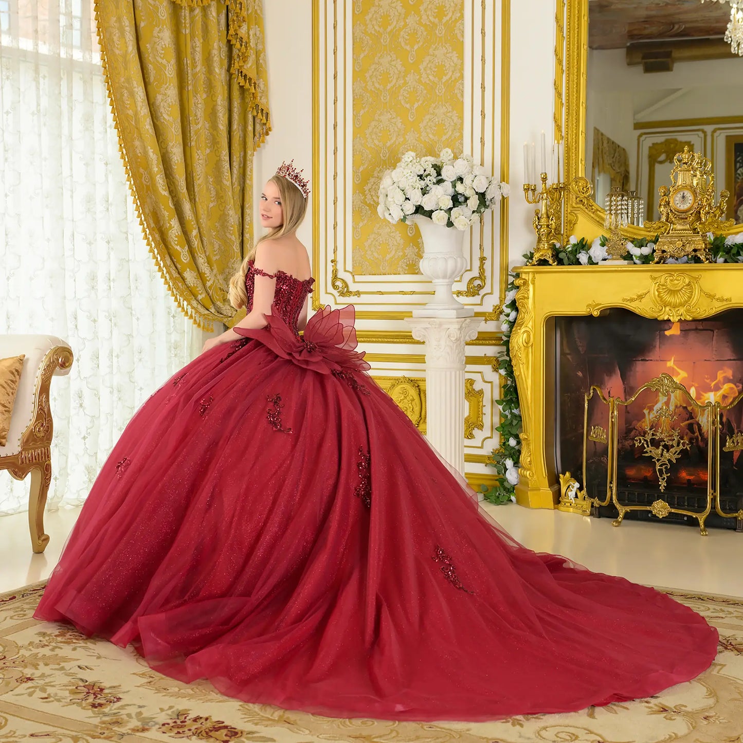 Girl wearing a burgundy quinceañera dress with tiara in an opulent room with gold decor and a fireplace.