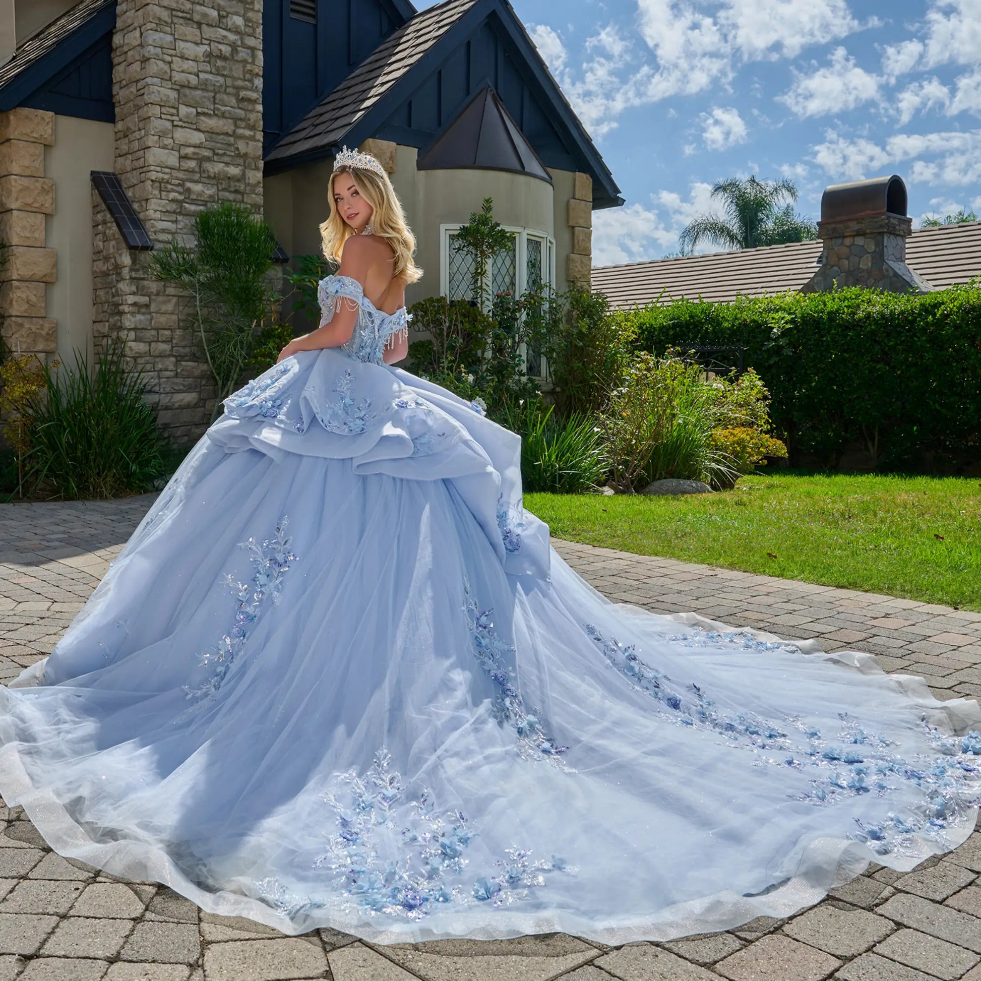 Wide back view of light blue quinceañera ball gown showing extended tulle train, layered skirt structure, floral appliqués, and outdoor garden backdrop