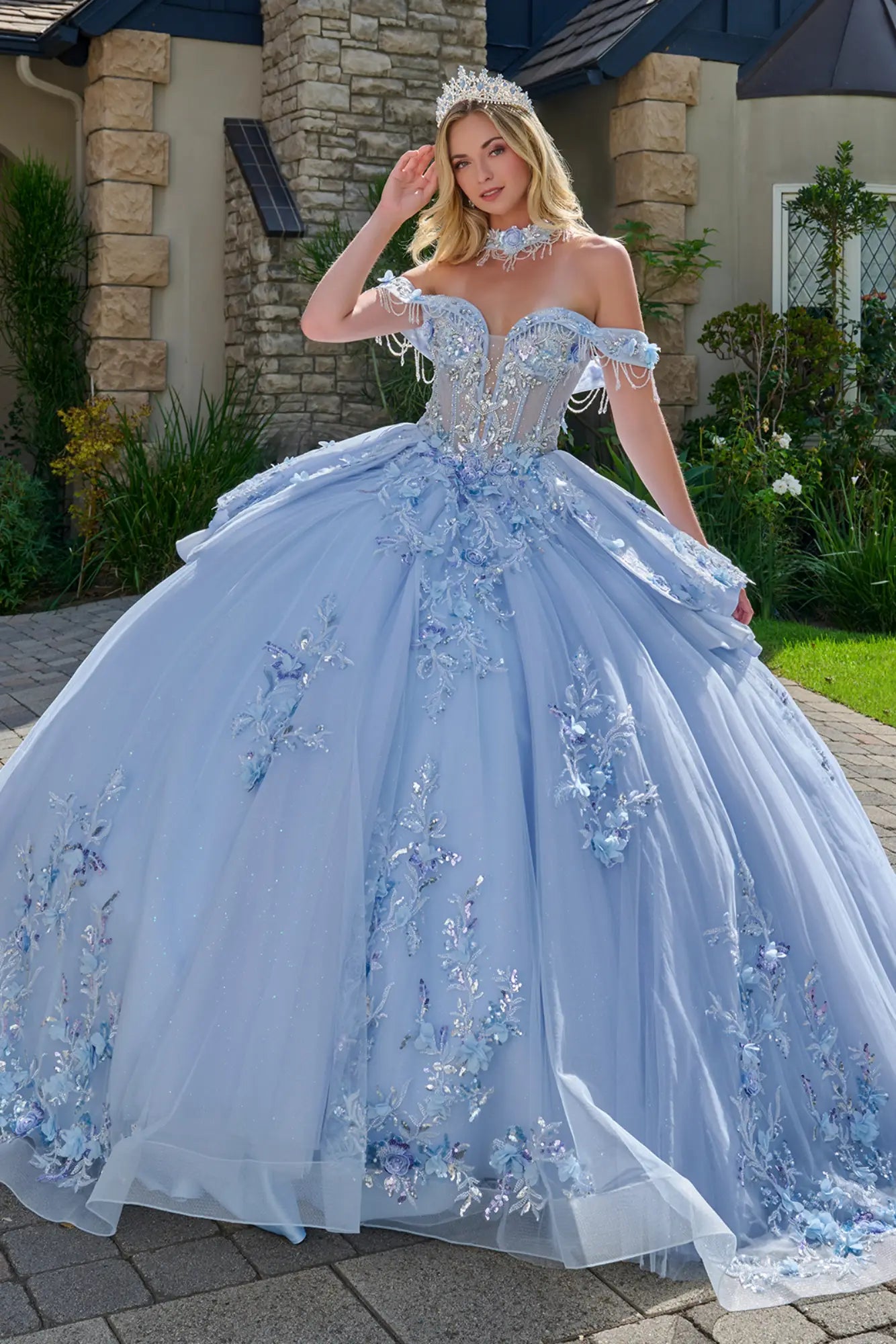 Front view of light blue quinceañera ball gown with off-the-shoulder sweetheart neckline, crystal-embellished corset bodice, floral appliqués, and full glitter tulle skirt in an outdoor estate setting