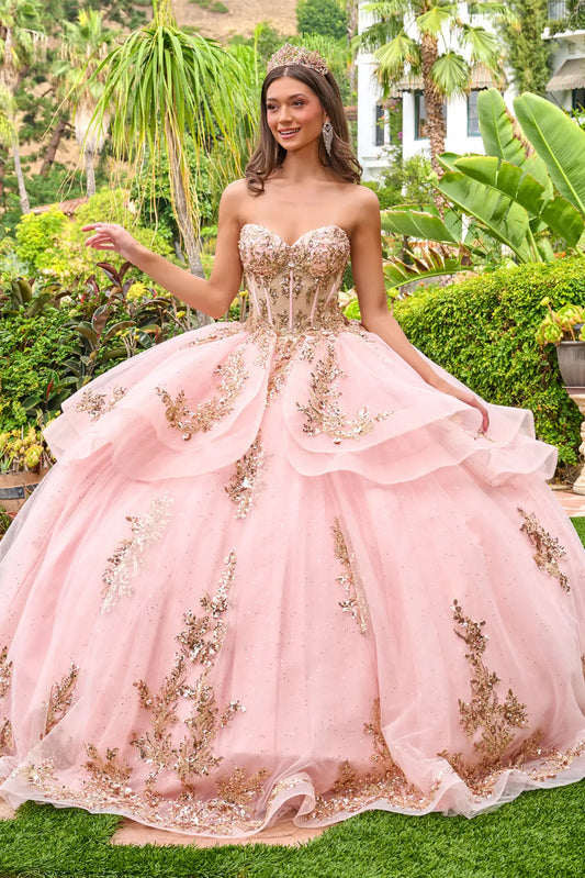 Front view of blush pink quinceañera ball gown with strapless corset bodice, gold embroidery, layered tulle skirt, and sparkling appliqués in a garden setting