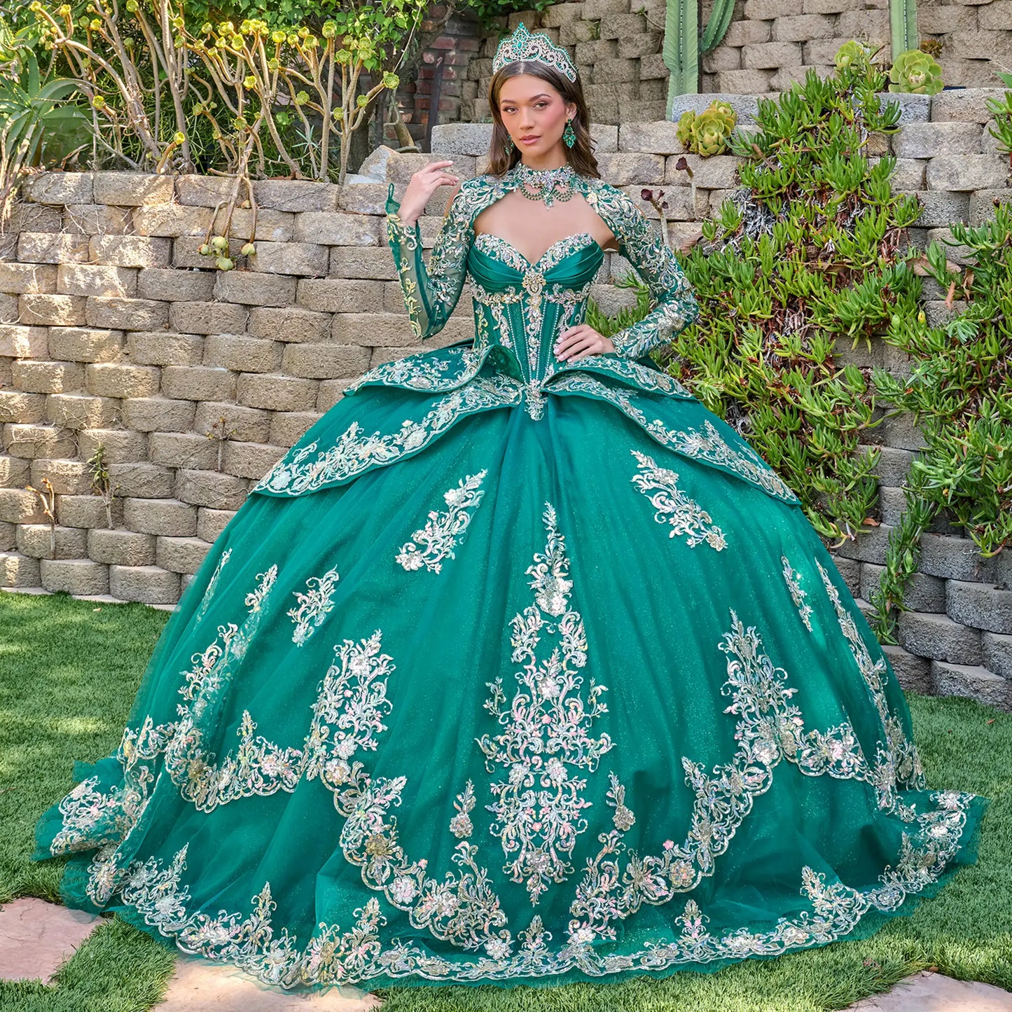 Woman in a green ball gown with silver embroidery standing outdoors against a stone wall.