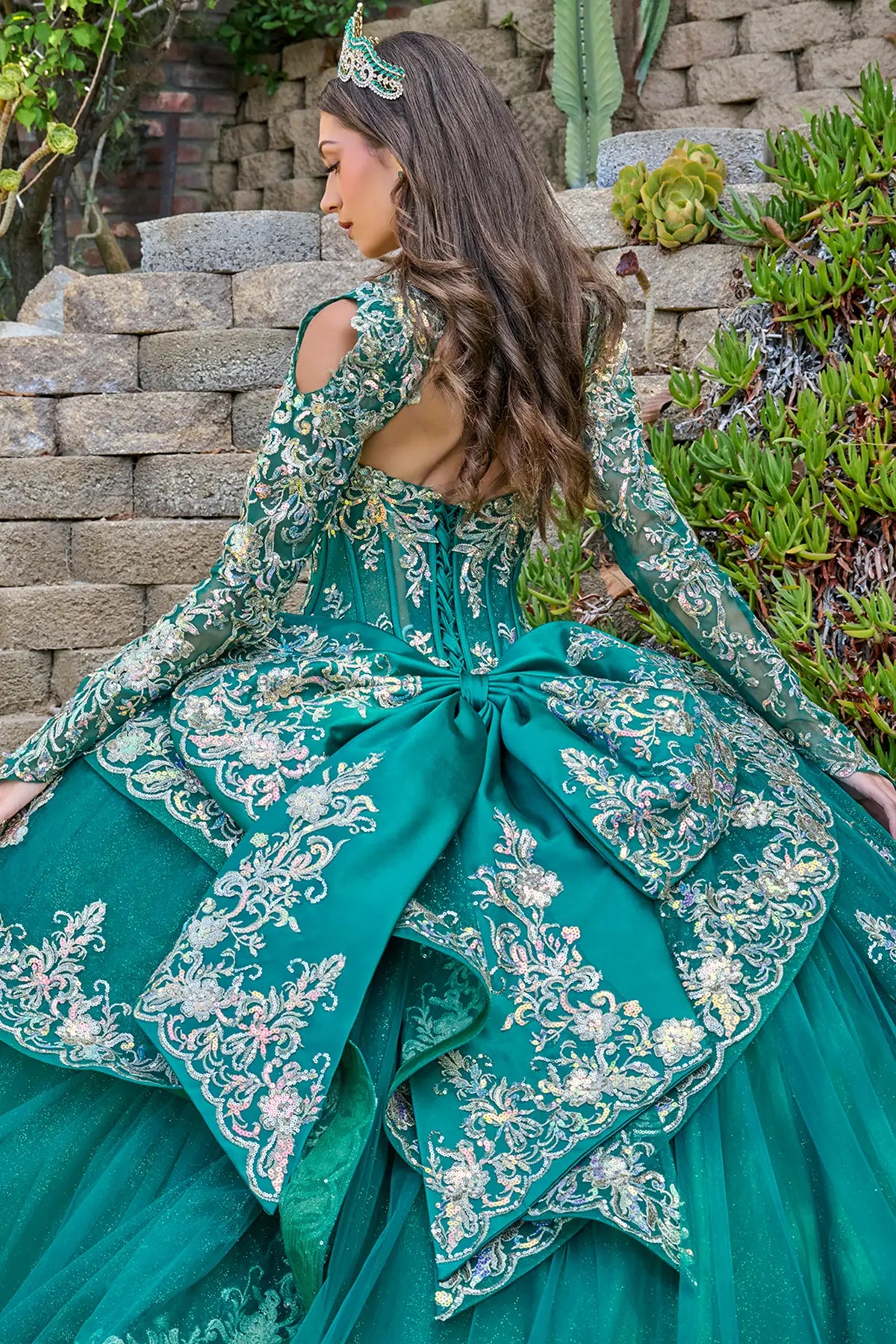 Woman in a green embroidered dress sitting outdoors with greenery and stone steps in the background.
