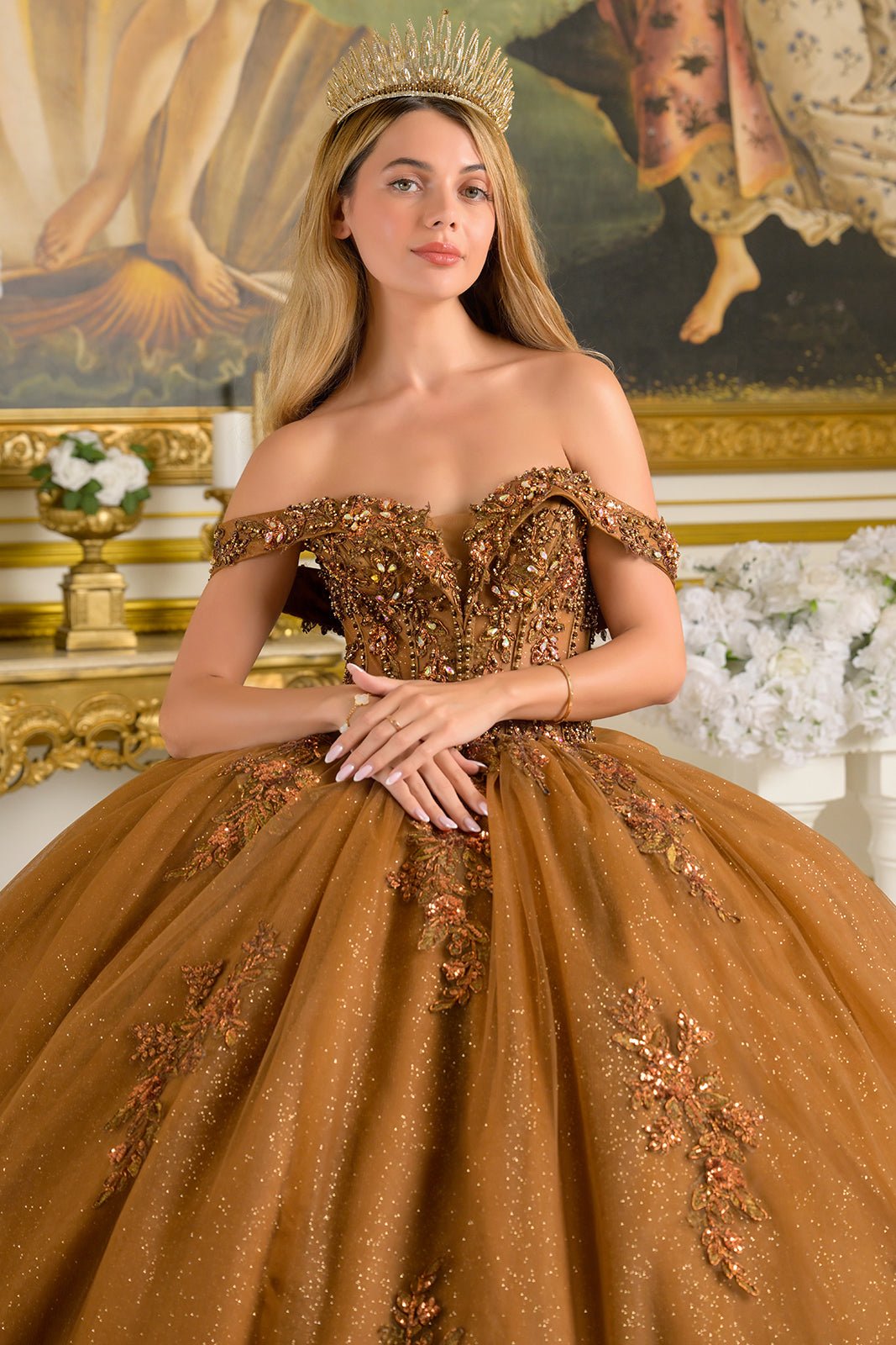 Woman wearing brown ball gown with floral embellishments, sparkling details, and crown in opulent room with gold accents and classical painting