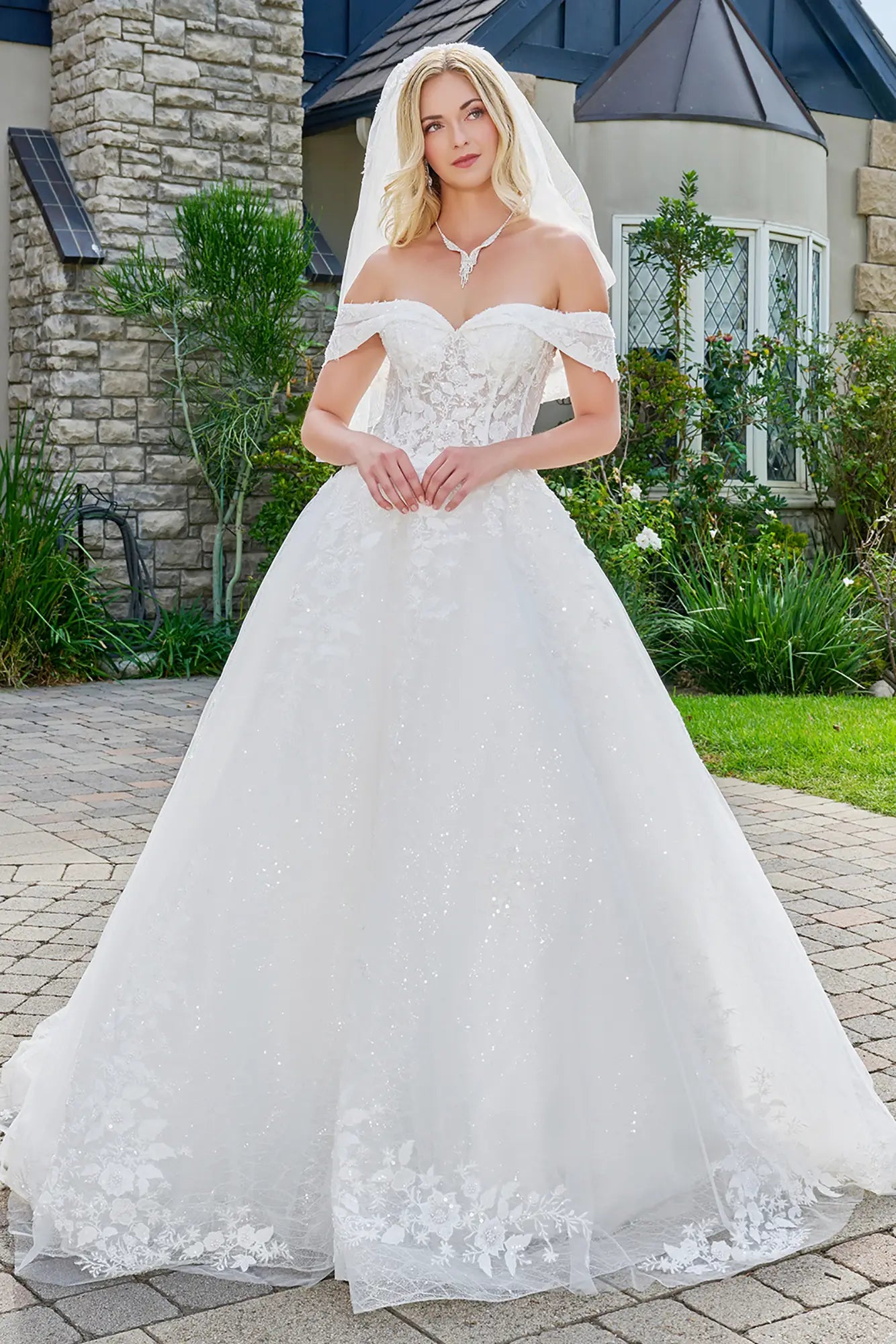 Woman in a white wedding dress standing outdoors with a stone building and garden in the background