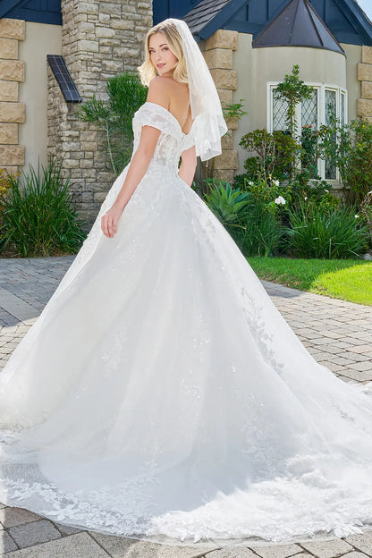 Woman in a white wedding dress standing outdoors with a stone building and garden in the background.