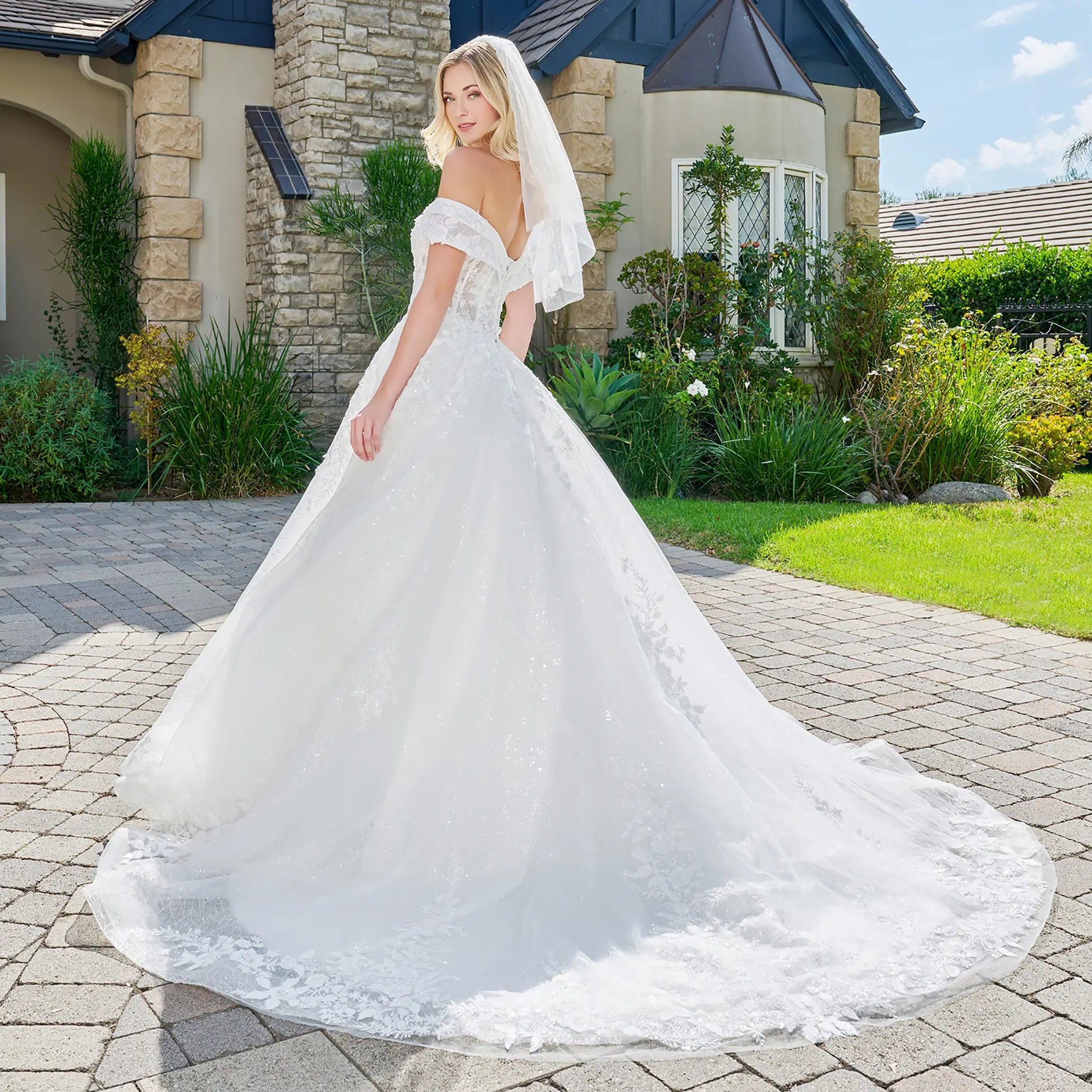Woman in a white wedding dress standing on a driveway with a house and garden in the background