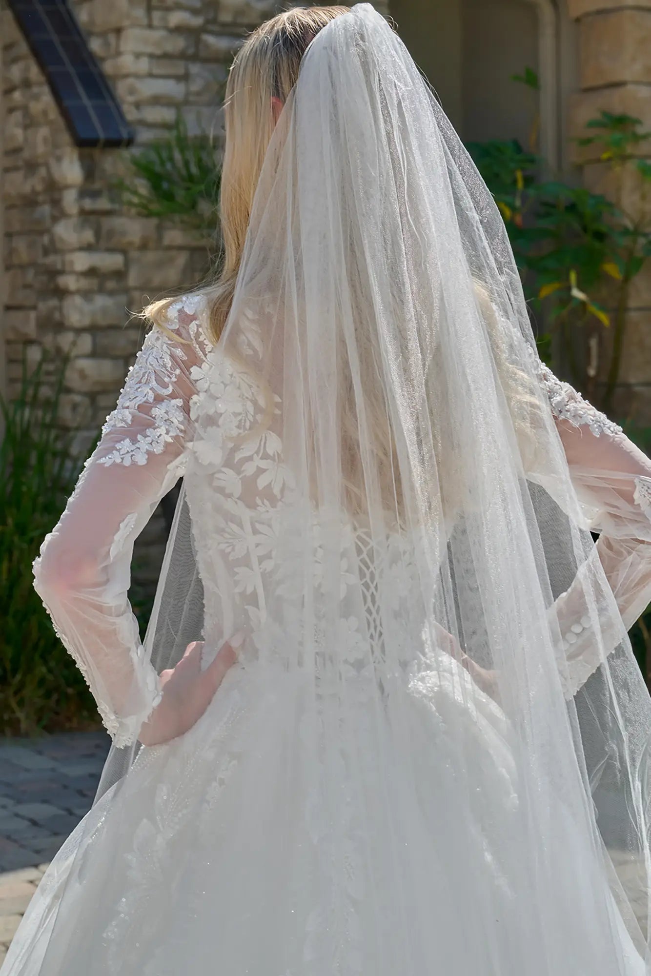 Person wearing a white lace wedding dress with a long veil outdoors.