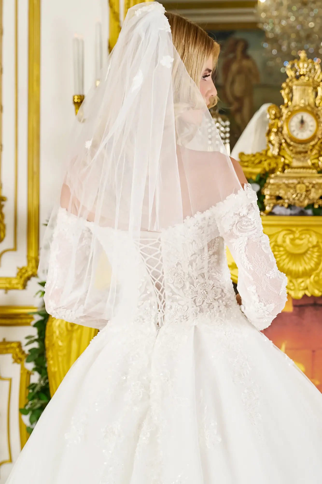 Person wearing a white wedding dress with a long train and veil in an ornate room.