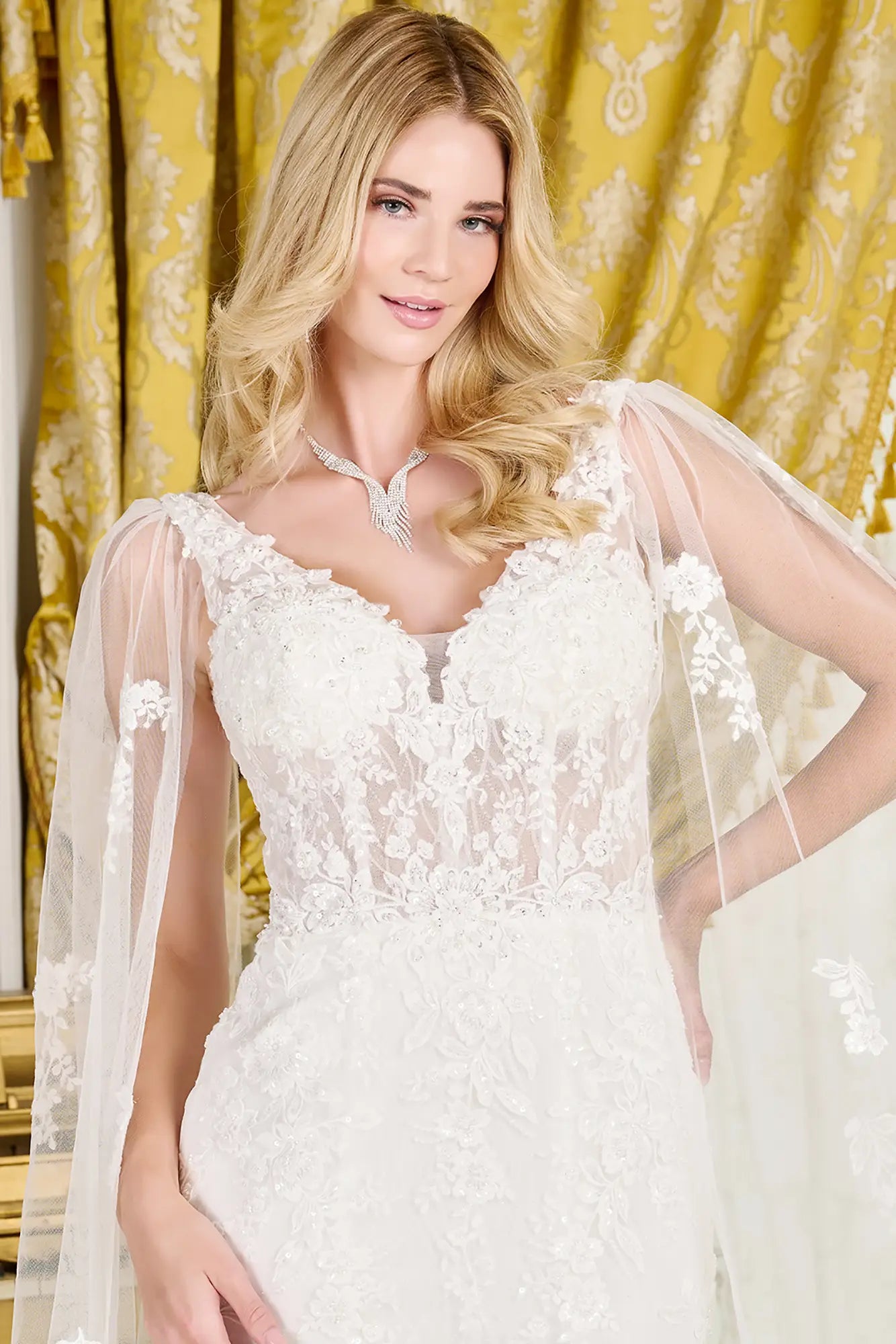 Woman wearing a white lace wedding dress with a sheer cape against a yellow curtain background