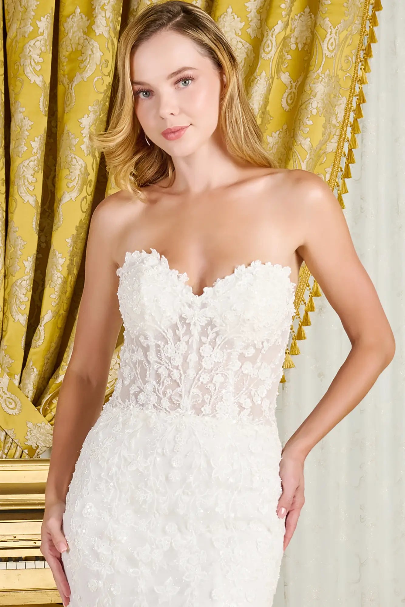 Woman wearing a strapless white lace wedding dress in front of a decorative yellow curtain.