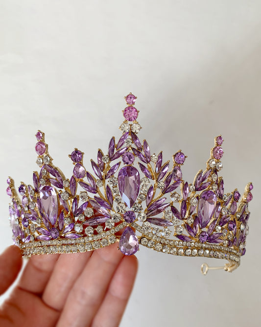 Purple and gold tiara held by a hand against a plain background
