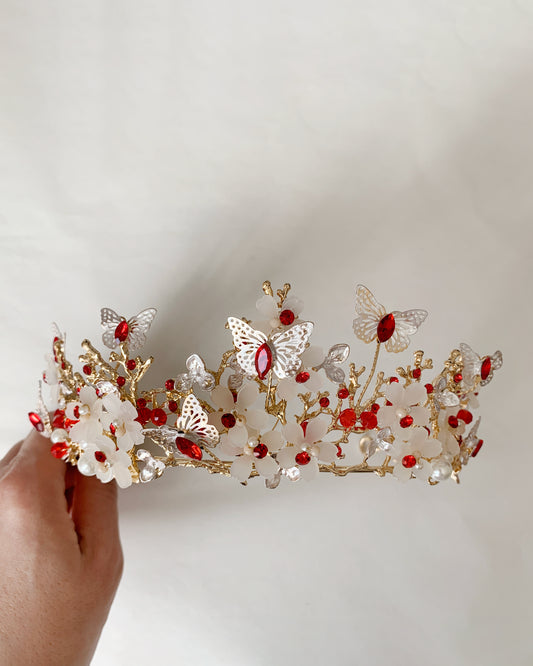 Decorative tiara with butterflies and red stones held by a hand against a plain background