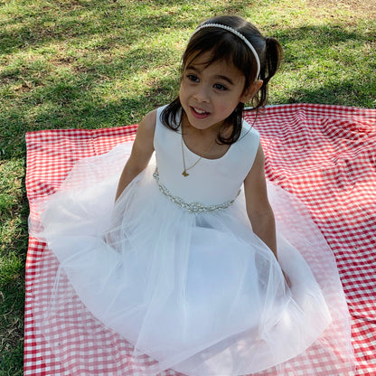 Young girl in a white dress sitting on a red and white checkered blanket in a grassy area.