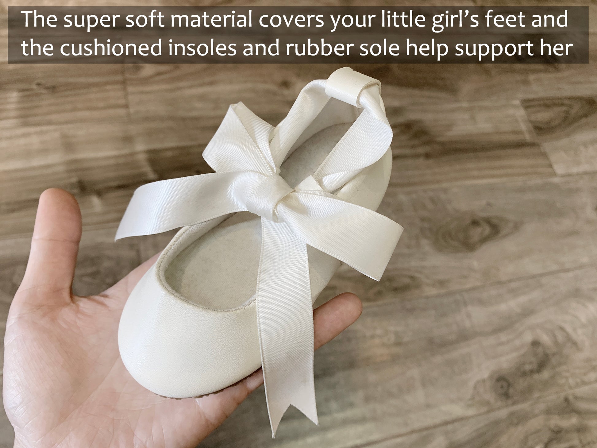 White ballerina shoe with a bow held by a hand on a wooden floor.