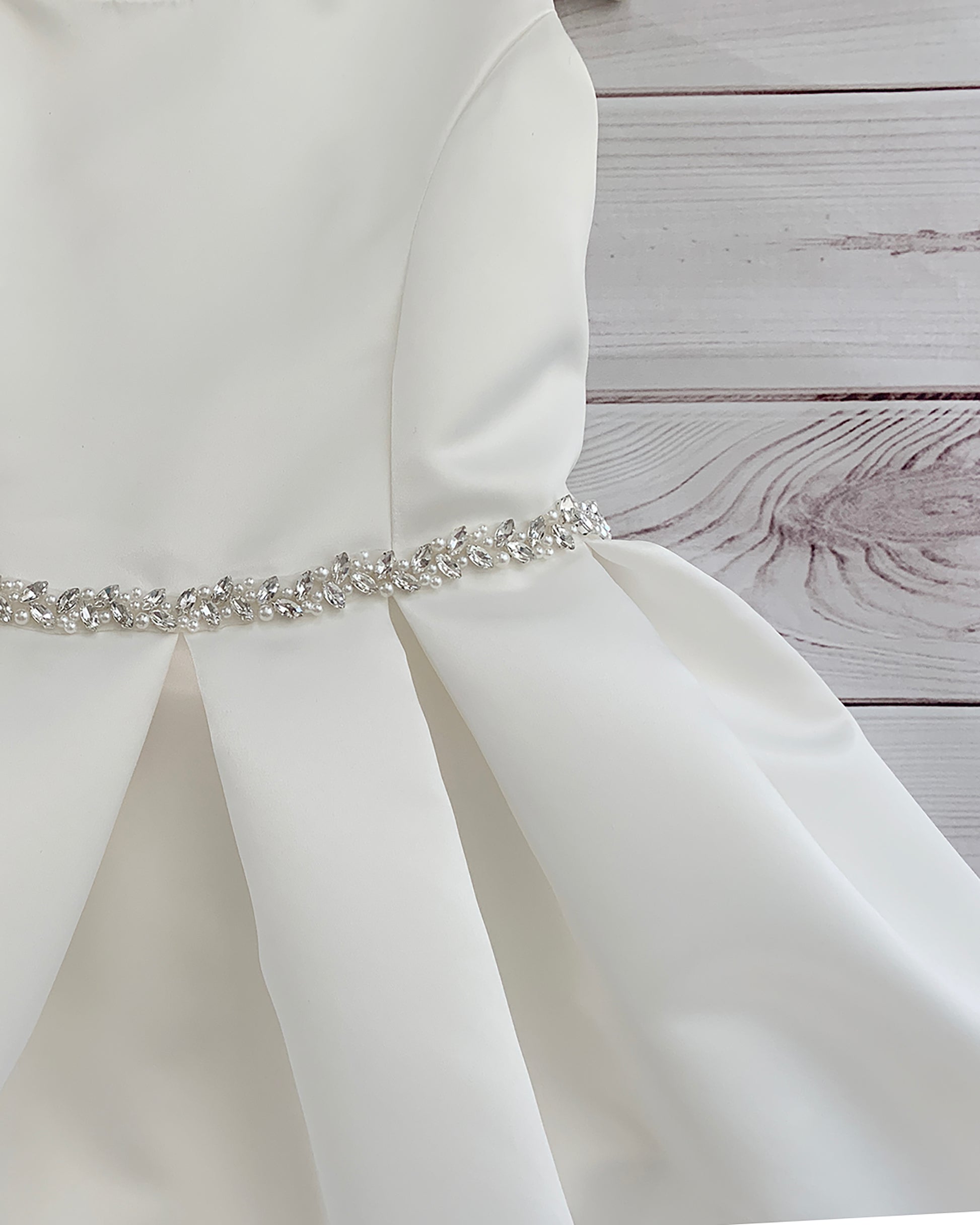 White satin flower girl dress with a decorative belt on a wooden surface.