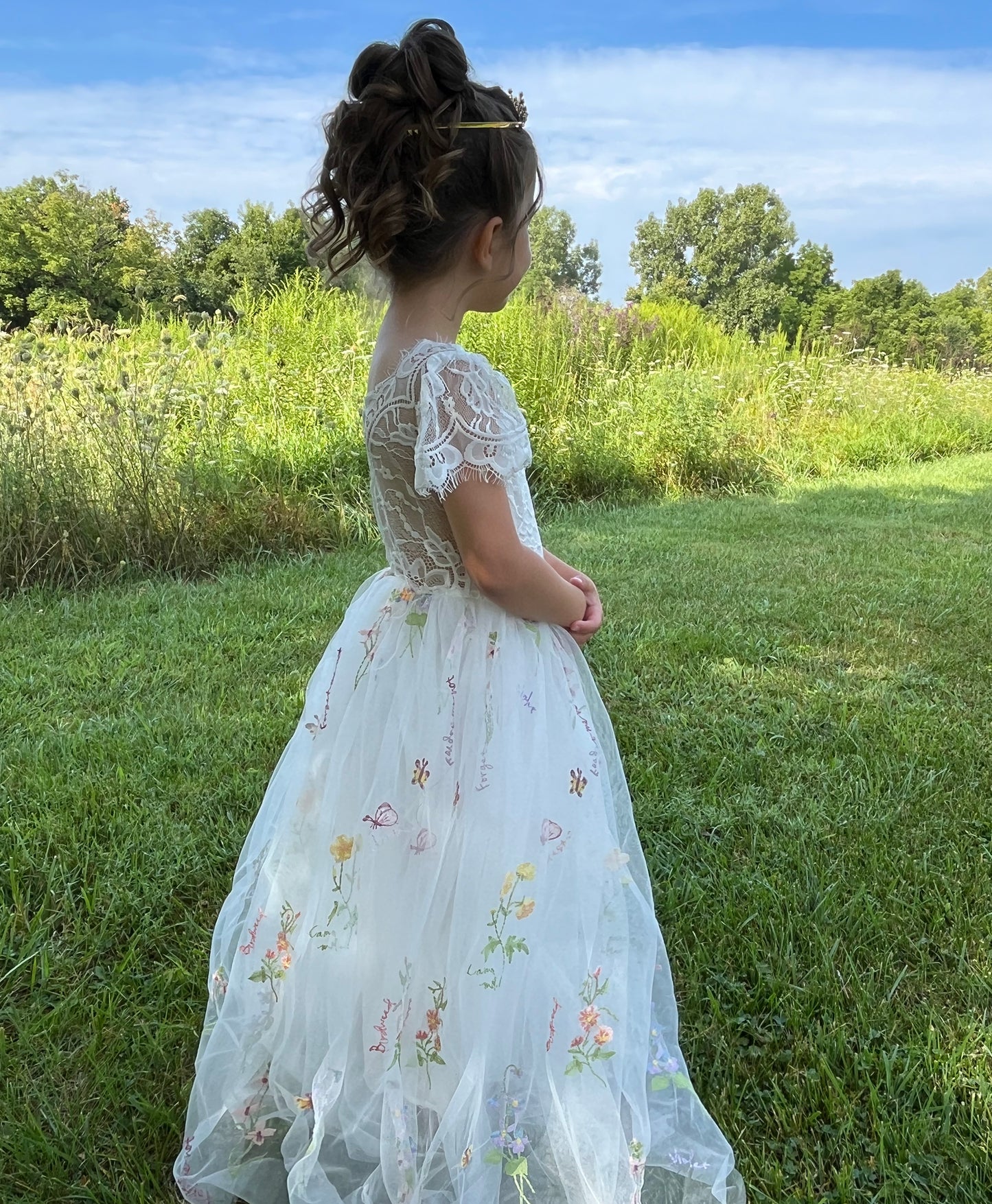 Young girl in a white floral dress standing in a grassy field with trees in the background
