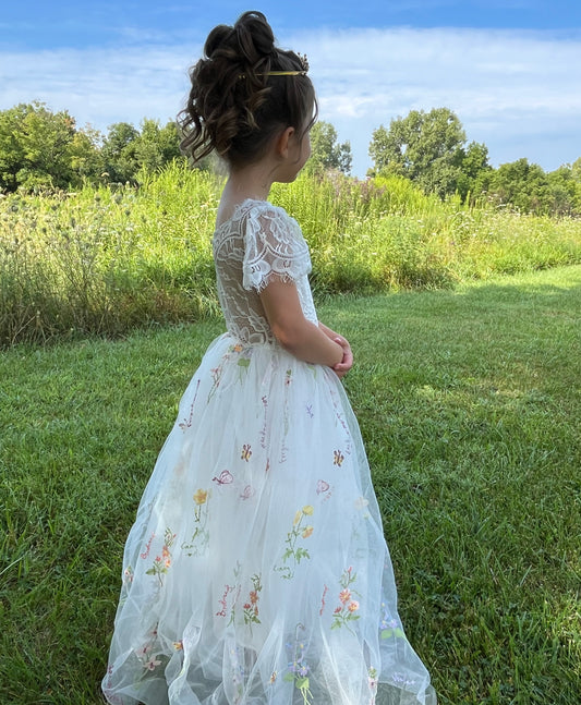 Young girl in a white floral dress standing in a grassy field with trees in the background