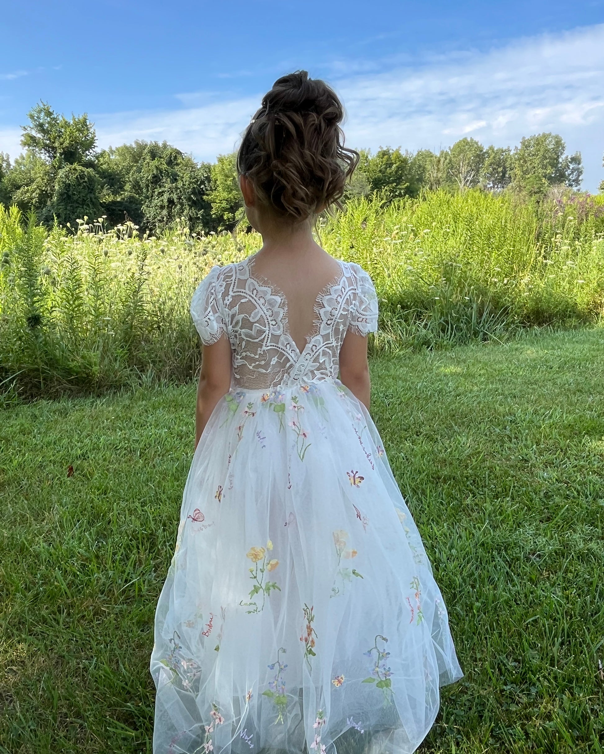 Young girl in a white lace dress with floral embroidery standing in a grassy field.