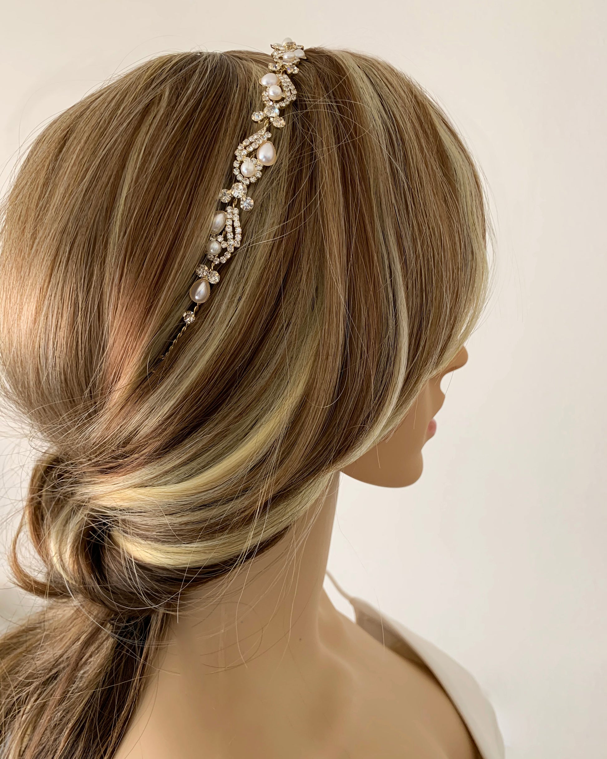 Mannequin head with blonde hair wearing a decorative gold pearl with rhinestones headband against a plain background
