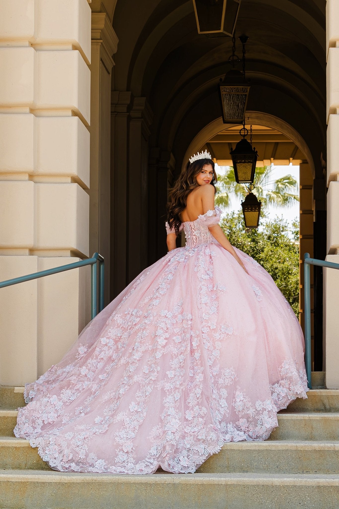 Woman in a pink ball gown with extra long train standing on steps with a building and greenery in the background