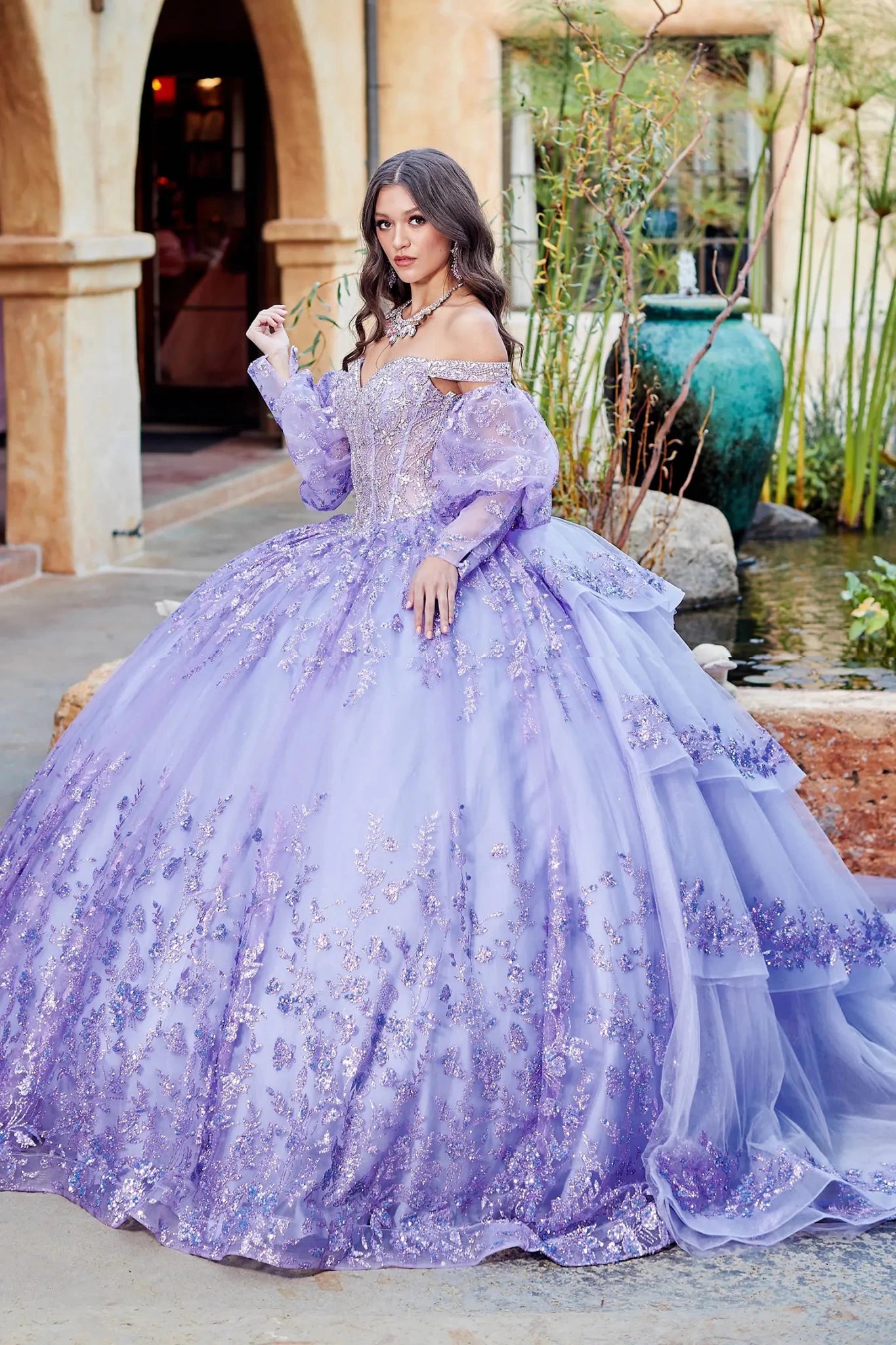 Woman in a lilac ball gown with long ruffled train and detachable sheer long sleeves standing outdoors with decorative elements.