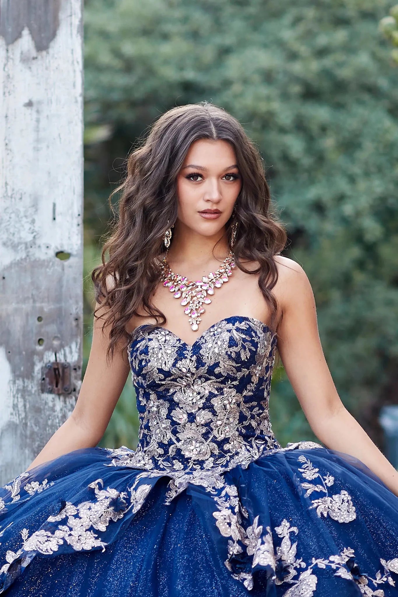 Woman in a blue and silver dress with jewelry, standing against a blurred natural background