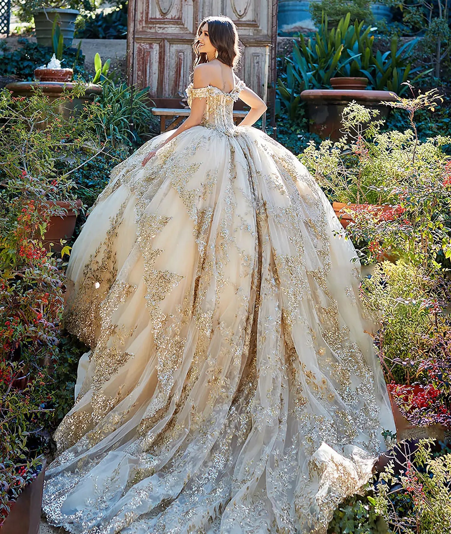 Girl in a sparkling off the shoulder gold champagne quinceañera dress with extra long train standing in a garden setting
