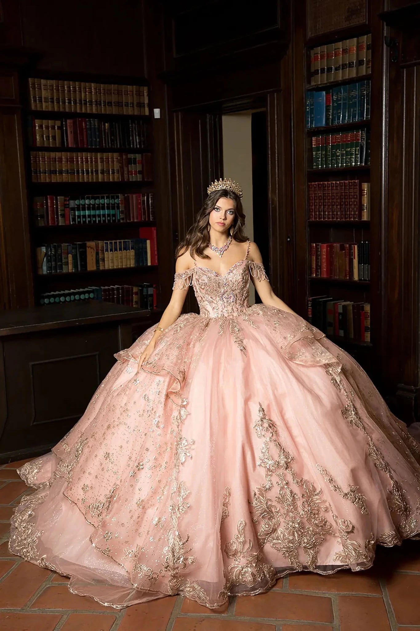 A girl in a rose gold quinceanera ball gown with intricate details standing in a room with bookshelves.