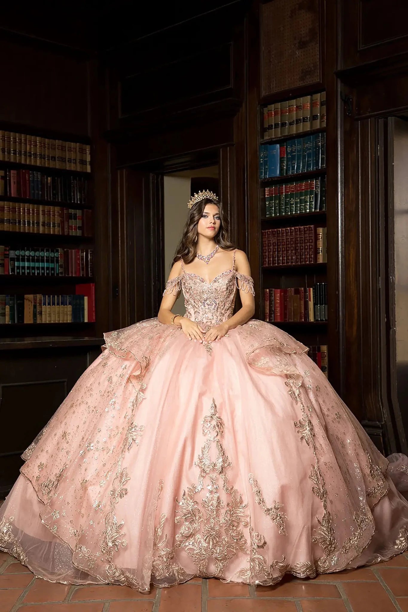 A girl in a rose gold ball gown with gold embroidery standing in a library.