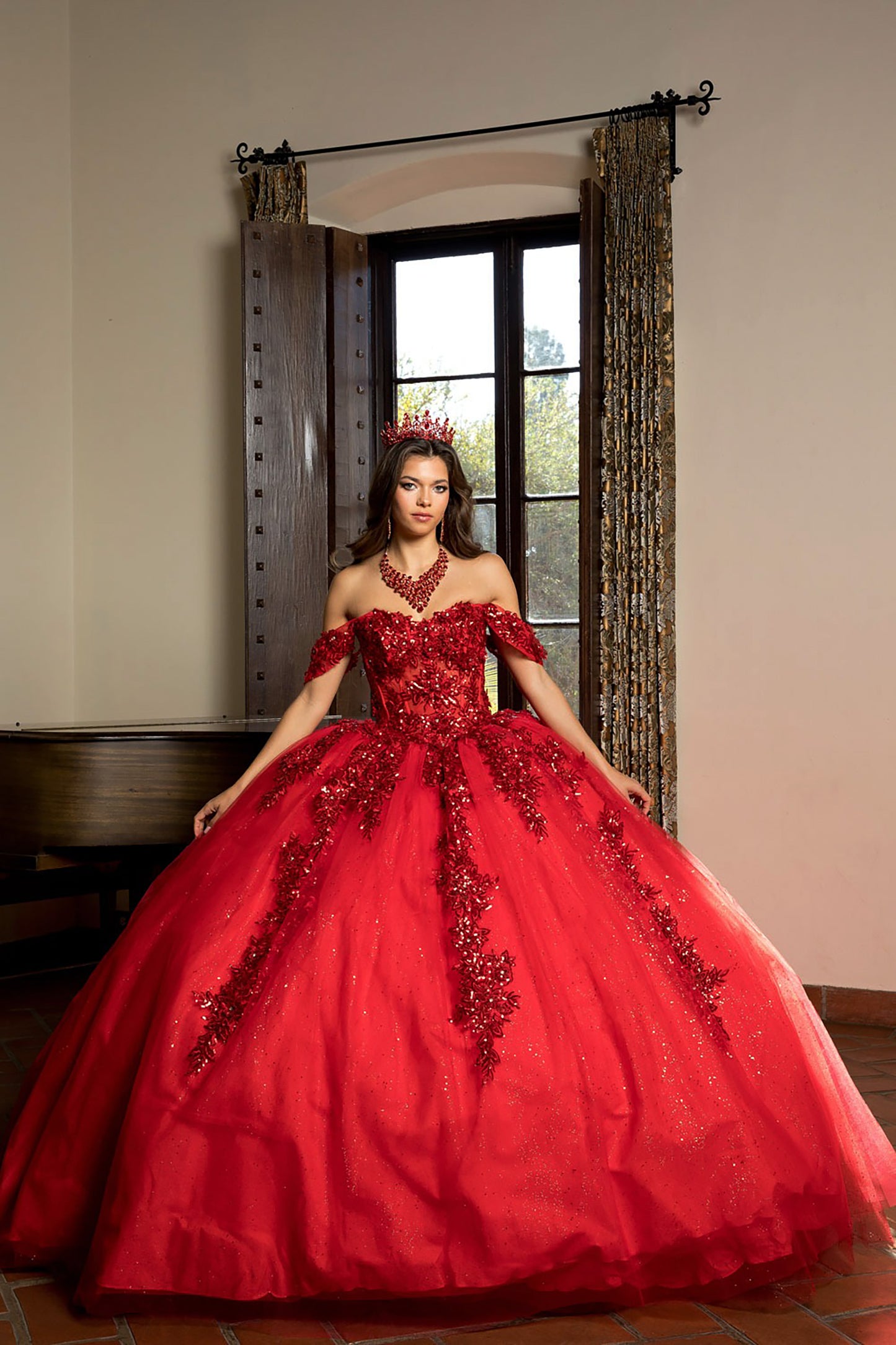 Woman in a red ball gown with floral details standing in a room with a window and curtains.