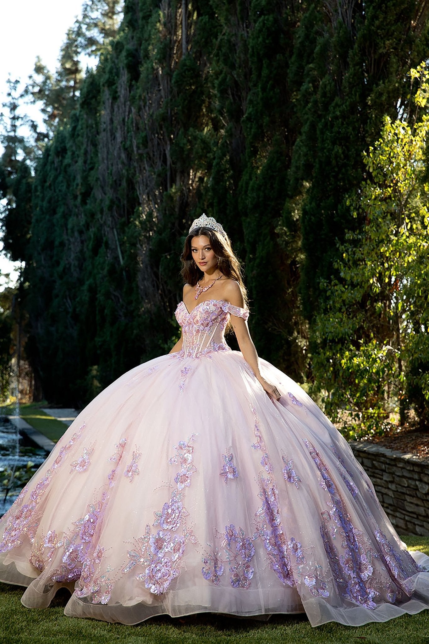 woman standing outdoors on a stone pathway beside a wall and tall trees, dressed in a dramatic light pink ball gown. The gown is voluminous, with a fitted bodice and off‑the‑shoulder sleeves, and it is richly decorated with intricate purple floral embellishments that cascade across the fabric