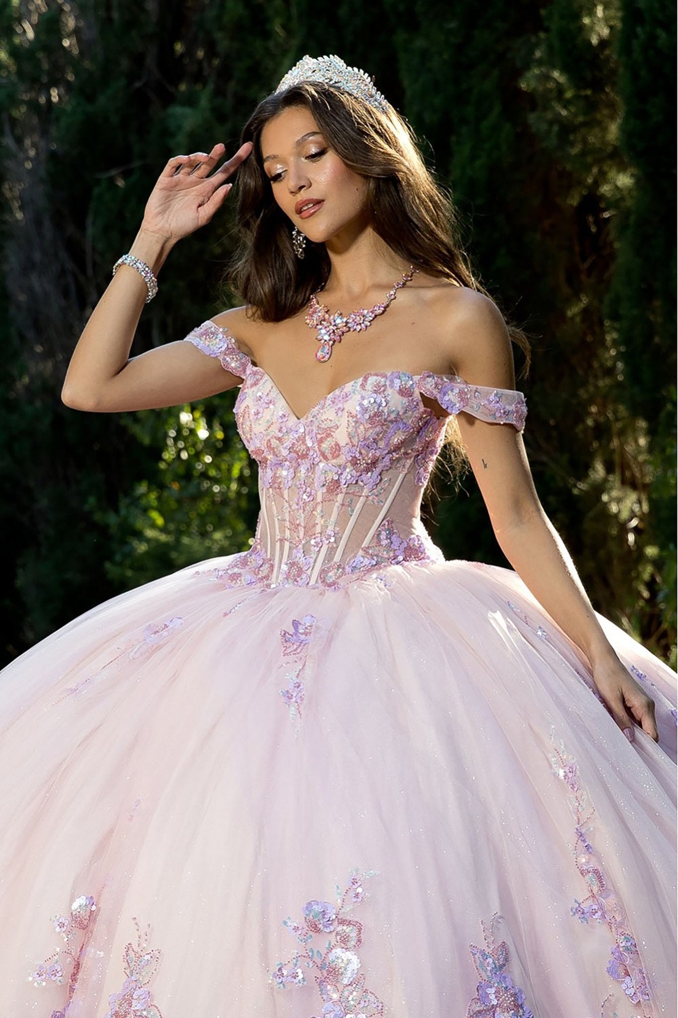 woman standing outdoors against a backdrop of lush greenery, dressed in an elaborate light pink ball gown. The gown features a fitted corset‑style bodice with off‑the‑shoulder sleeves and a voluminous skirt adorned with delicate purple and pink floral appliqués.