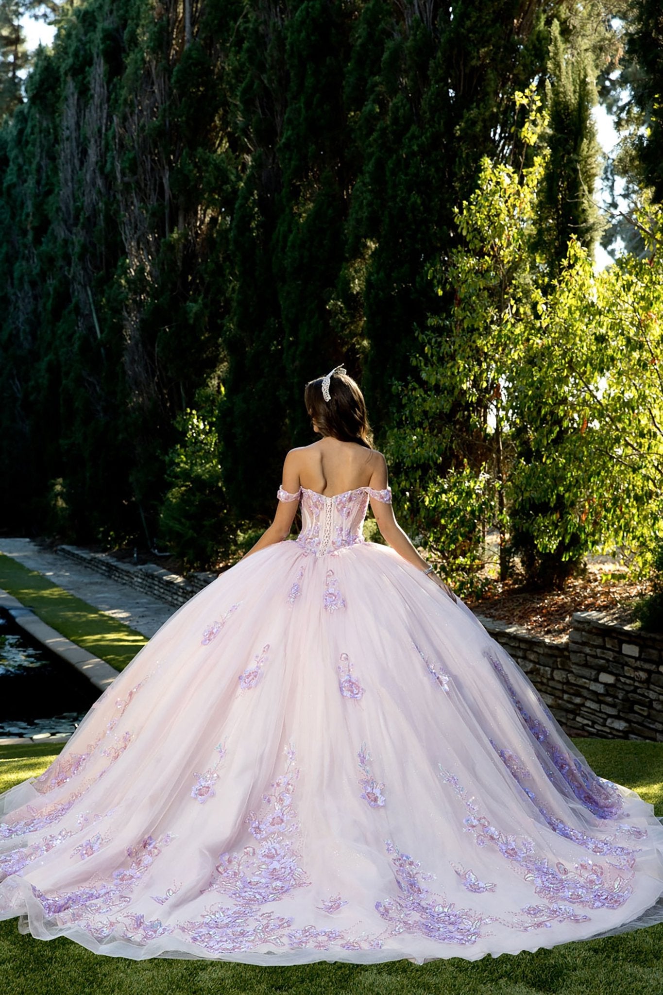 woman standing outdoors on a manicured lawn, surrounded by tall trees and a stone wall. She is wearing an elaborate floor‑length ball gown in a soft light pink or lavender tone, richly decorated with intricate floral embellishments and embroidery concentrated along the lower portion of the skirt.