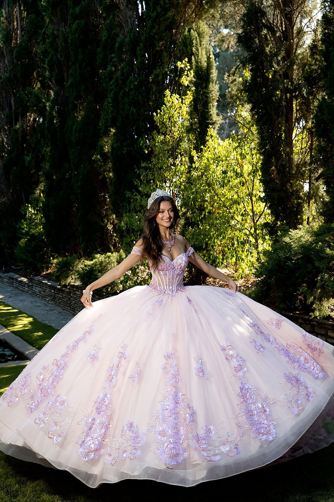 A girl wearing a blush pink quinceañera dress with floral sequins and a sweet 15 design, standing and smiling in an outdoor setting with trees in the background.